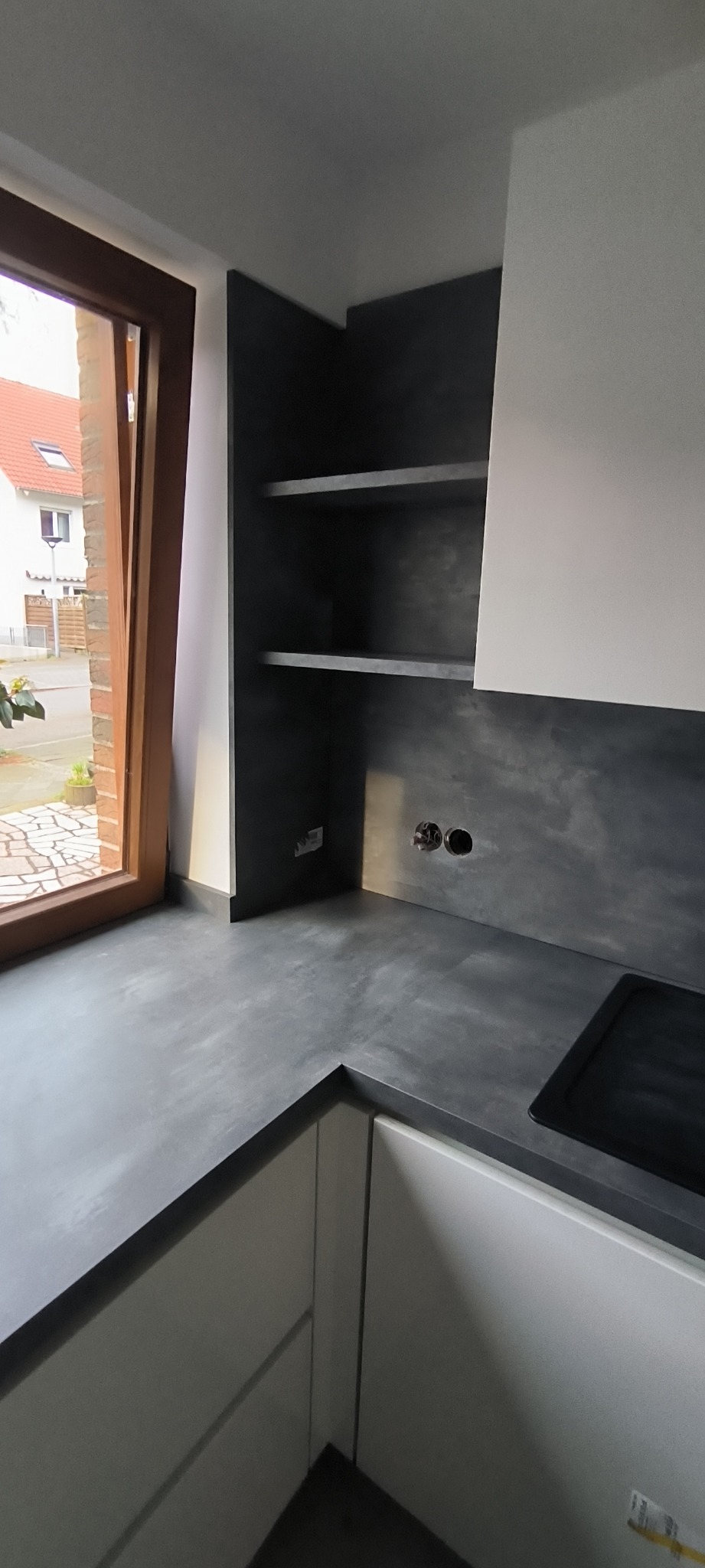 Nowoczesna kuchnia z szarą, teksturowaną ścianą i półkami, widoczne puszki elektryczne, fragment blatu i okno z widokiem na budynki.