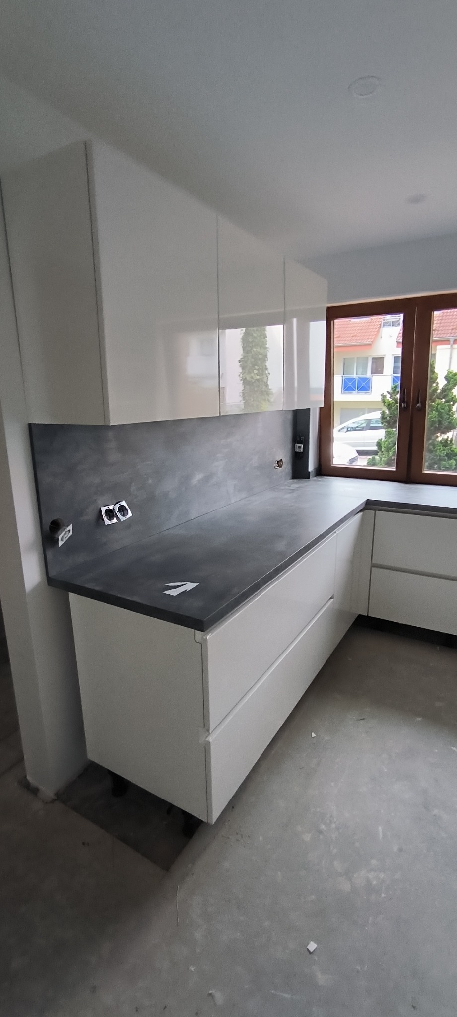 Nowoczesna kuchnia w trakcie wykańczania: białe szafki z szarym blatem i ścianą, widoczne gniazdka elektryczne, surowy beton na podłodze, brak cokołów dolnych szafek, okno z drewnianą ramą...