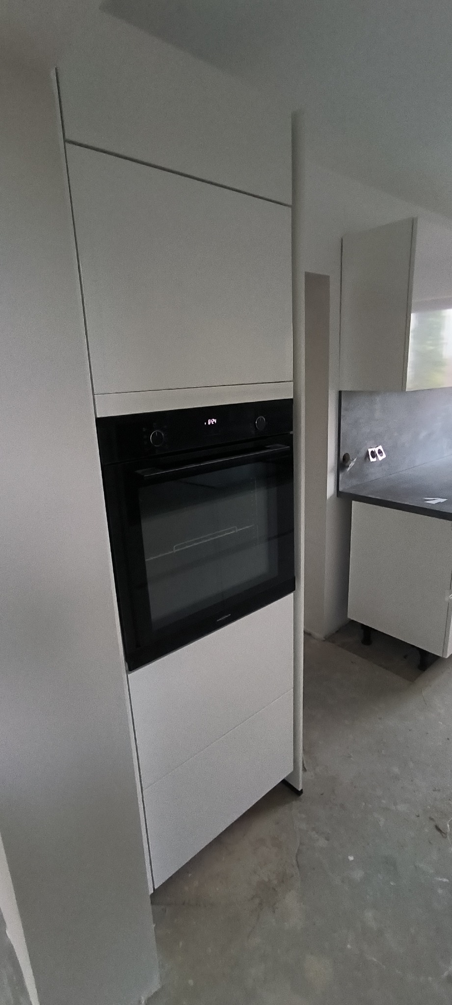 Wbudowany piekarnik w białej zabudowie kuchennej, widoczny fragment blatu i podłoga w trakcie remontu.