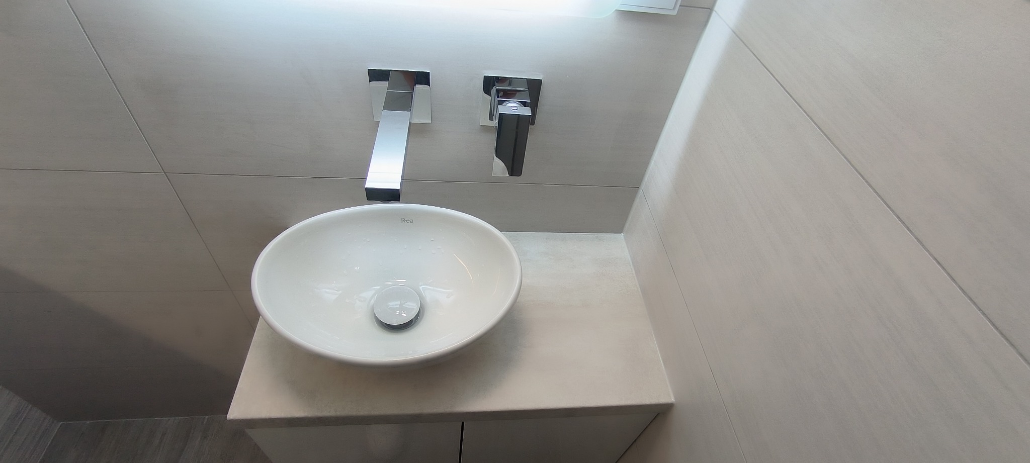 Nowoczesna, owalna umywalka marki Rea z chromowaną baterią ścienną, zamontowana na blacie z szarej płyty, otoczona jasnoszarymi płytkami ściennymi, oświetlona górnym światłem.
