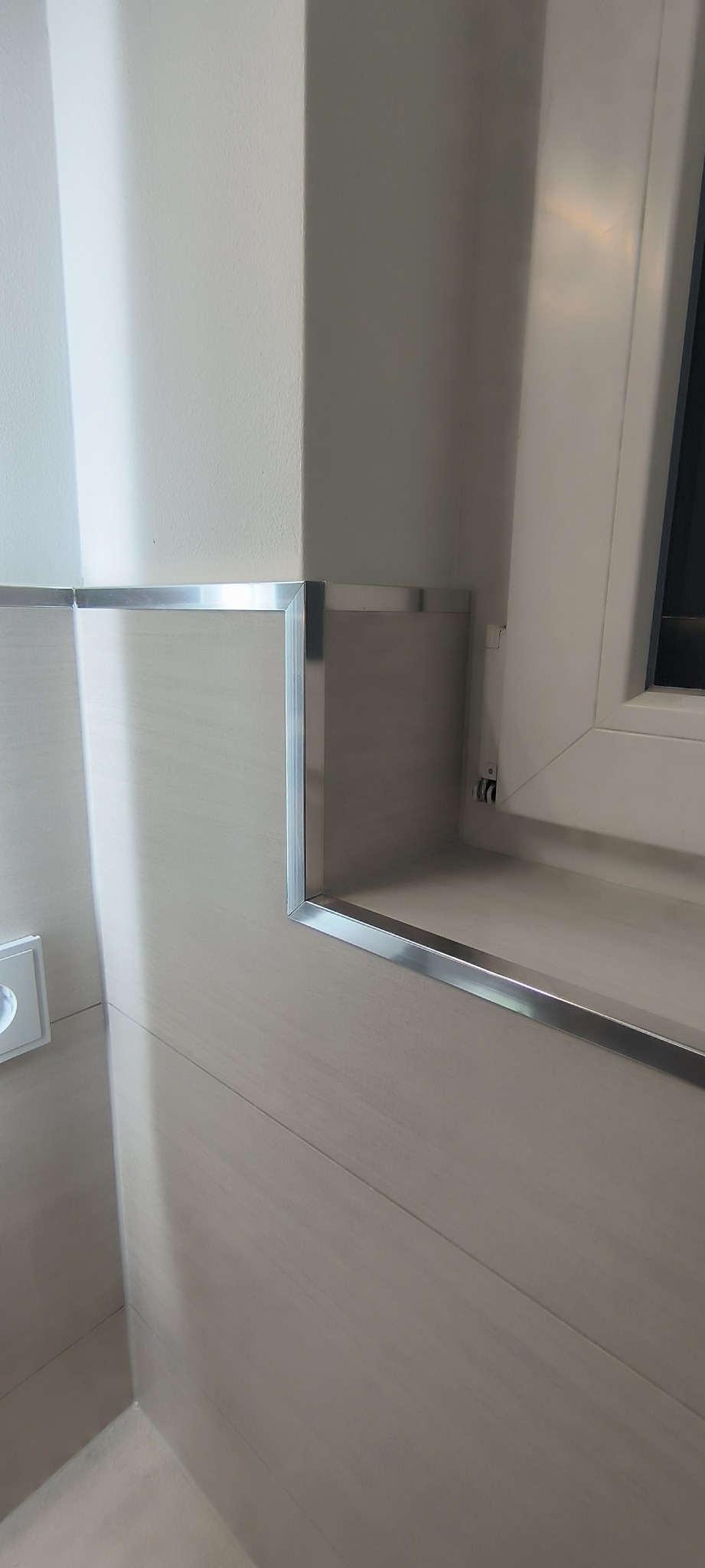 Wykończenie ściany w łazience z beżowymi płytkami imitującymi drewno, wykończone aluminiowymi listwami przy oknie i w rogu, widoczny fragment gniazdka elektrycznego.