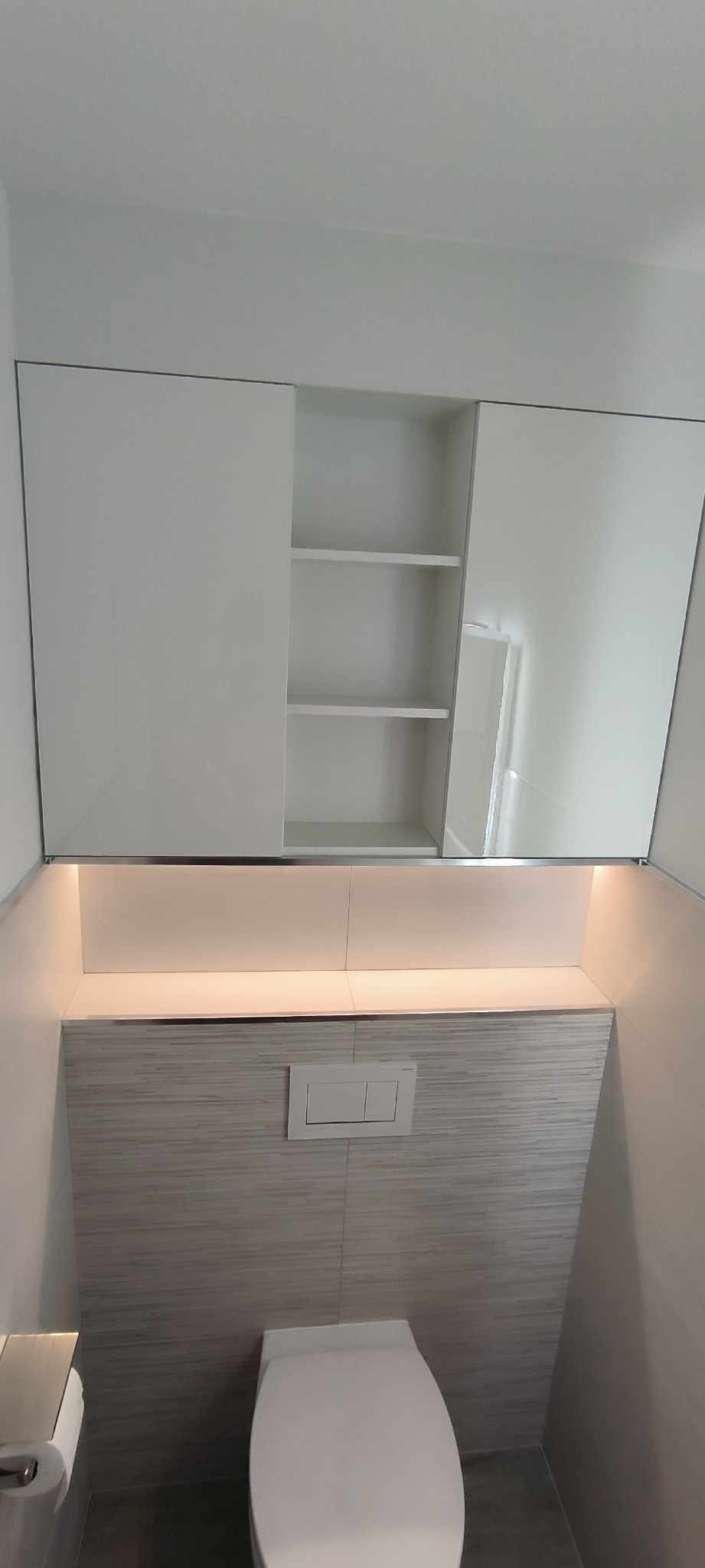 Nowoczesna toaleta w jasnych barwach z podświetlaną szafką nad zabudowaną spłuczką, wykończona płytkami imitującymi drewno