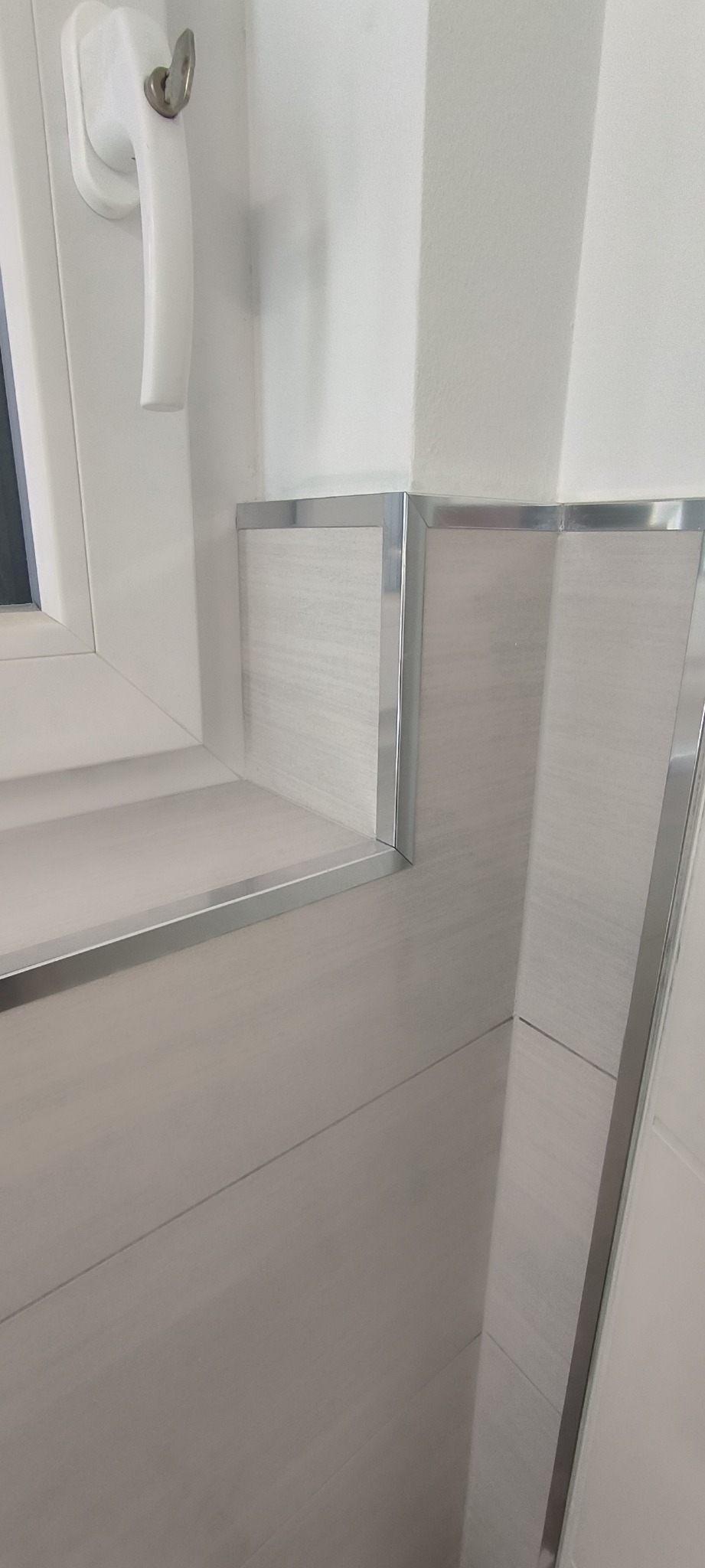 Wykończona narożna ściana w łazience z jasnymi płytkami imitującymi drewno i metalowymi listwami, obok okna z białą ramą.