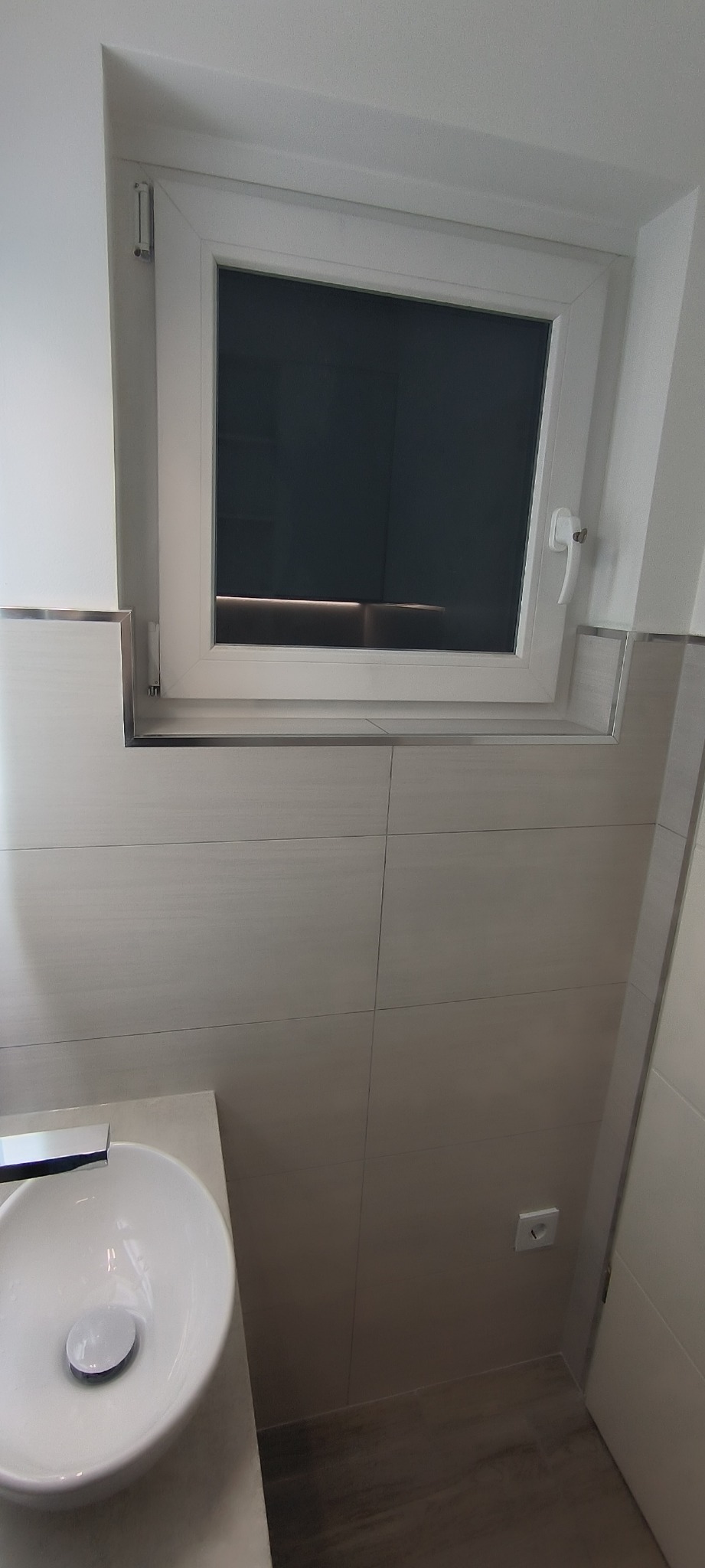 Wykończona łazienka z umywalką nablatową, dużymi płytkami imitującymi drewno i białym oknem PCV z chromowanymi listwami wykończeniowymi.