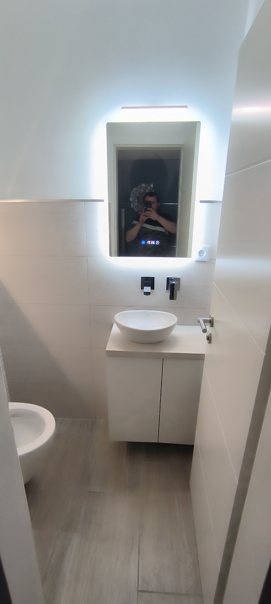 Nowoczesna łazienka z umywalką nablatową, podświetlanym lustrem z wyświetlaczem, minimalistyczną armaturą i płytkami imitującymi drewno na podłodze. W odbiciu lustra widoczna osoba robiąca zdjęcie.