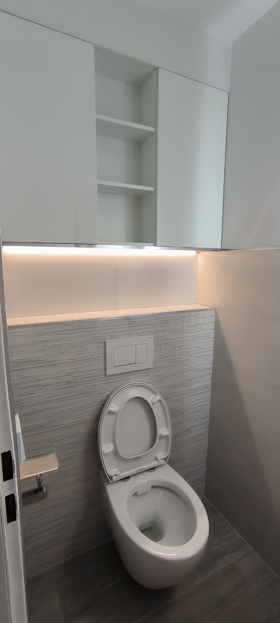 Nowoczesna toaleta wisząca z podświetleniem LED nad płytkami imitującymi drewno, zabudowa meblowa w kolorze białym z półkami.