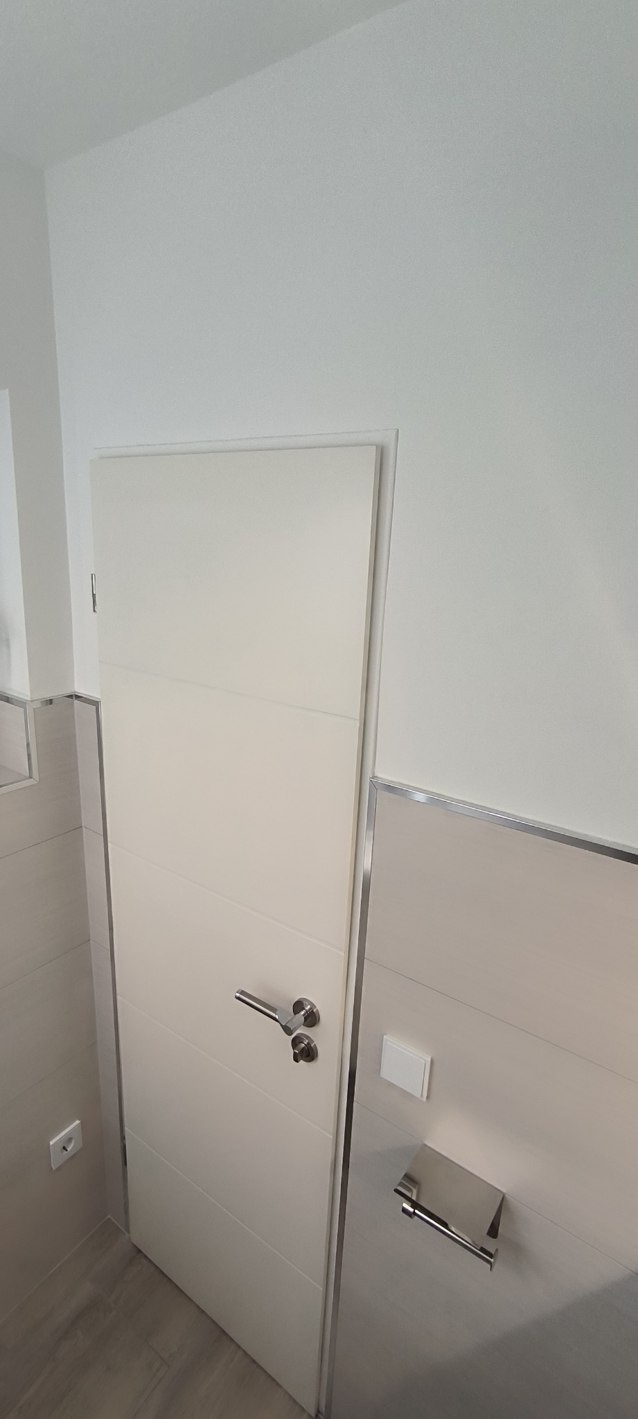 Nowoczesna, biała łazienka z płytkami imitującymi kamień, białą, minimalistyczną drzwiami z poziomymi frezowaniami i chromowanymi akcentami, takimi jak uchwyt na papier toaletowy i klamka.