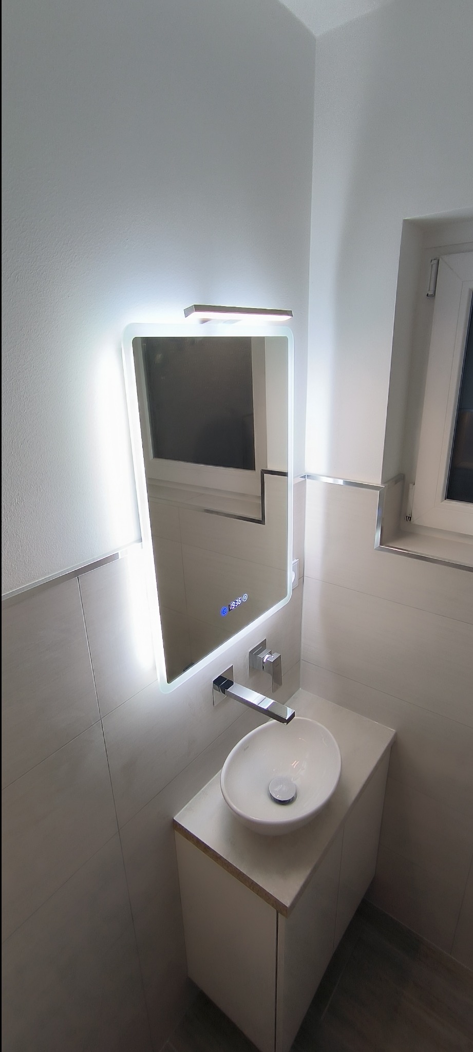 Nowoczesna łazienka z podświetlanym lustrem z wyświetlaczem cyfrowym, umywalką nablatową w kształcie misy i minimalistyczną baterią ścienną.