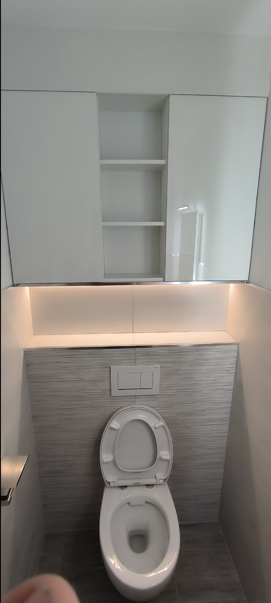 Nowoczesna toaleta podwieszana z zabudową, szara płytka imitująca drewno na ścianie, białe szafki z półkami nad toaletą, oświetlenie LED podszafkowe.