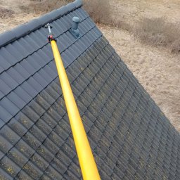 Dachy Cirocki - Czyszczenie ciemnoszarej dachówki z mchu za pomocą żółtej lancy teleskopowej. Widok z góry na dach i otoczenie z suchą trawą.