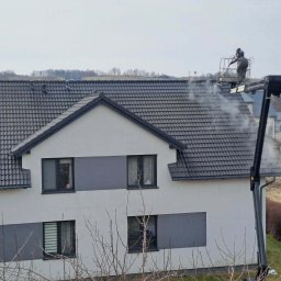 Dachy Cirocki - Pracownik na podnośniku czyści dach domu jednorodzinnego za pomocą myjki ciśnieniowej. Para wodna unosi się znad ciemnej dachówki. Widok z boku na budynek i okolicę.