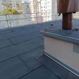 Dachy Cirocki - Płaski dach pokryty papą z brązowym kominem wentylacyjnym i zabezpieczeniami. W tle budynki mieszkalne i błękitne niebo. Widok z góry na fragment dachu.