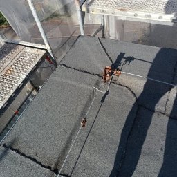 Dachy Cirocki - Szary dach z papy widziany z góry, z zamontowanym systemem odgromowym i fragmentem rusztowania. Widoczne łączenia papy i cień osoby wykonującej prace.