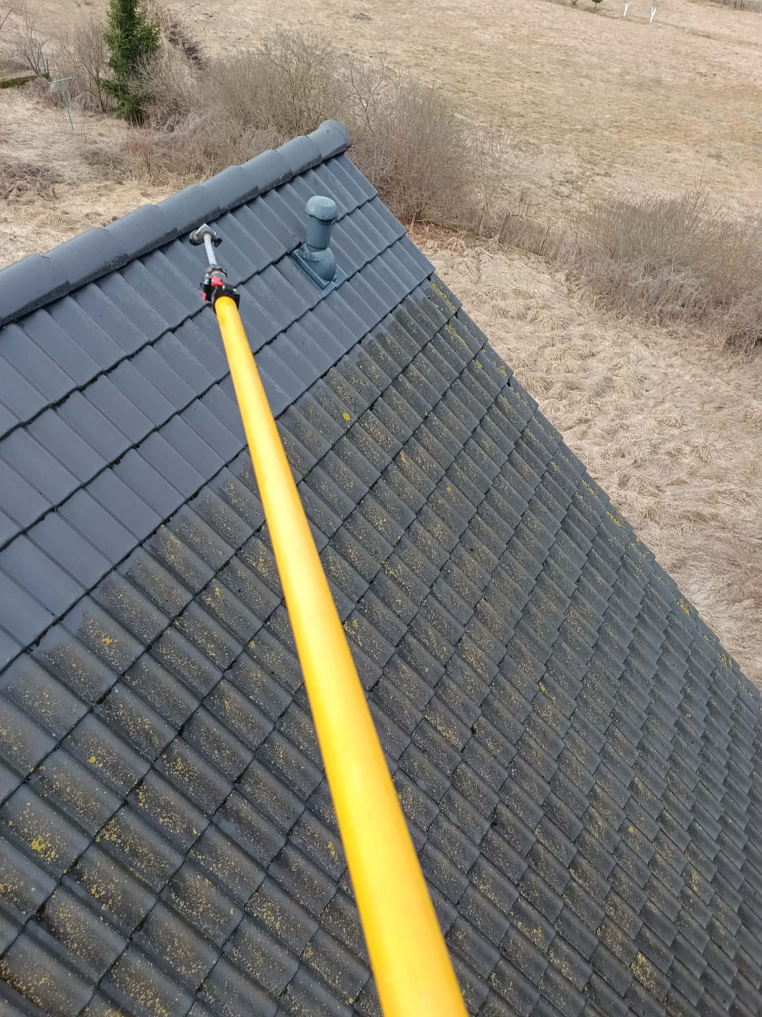 Czyszczenie ciemnoszarej dachówki z mchu za pomocą żółtej lancy teleskopowej. Widok z góry na dach i otoczenie z suchą trawą.