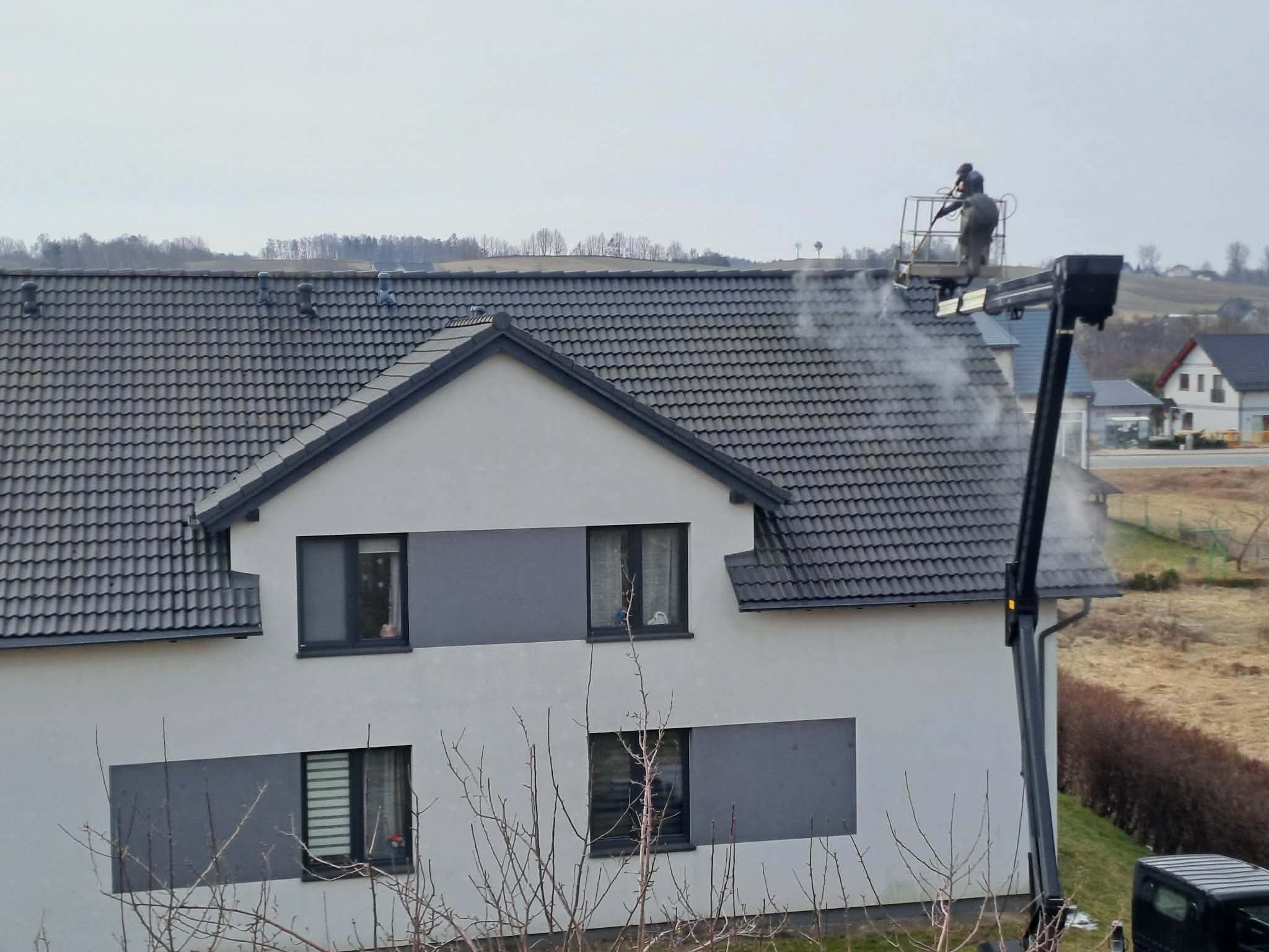 Pracownik na podnośniku czyści dach domu jednorodzinnego za pomocą myjki ciśnieniowej. Para wodna unosi się znad ciemnej dachówki. Widok z boku na budynek i okolicę.