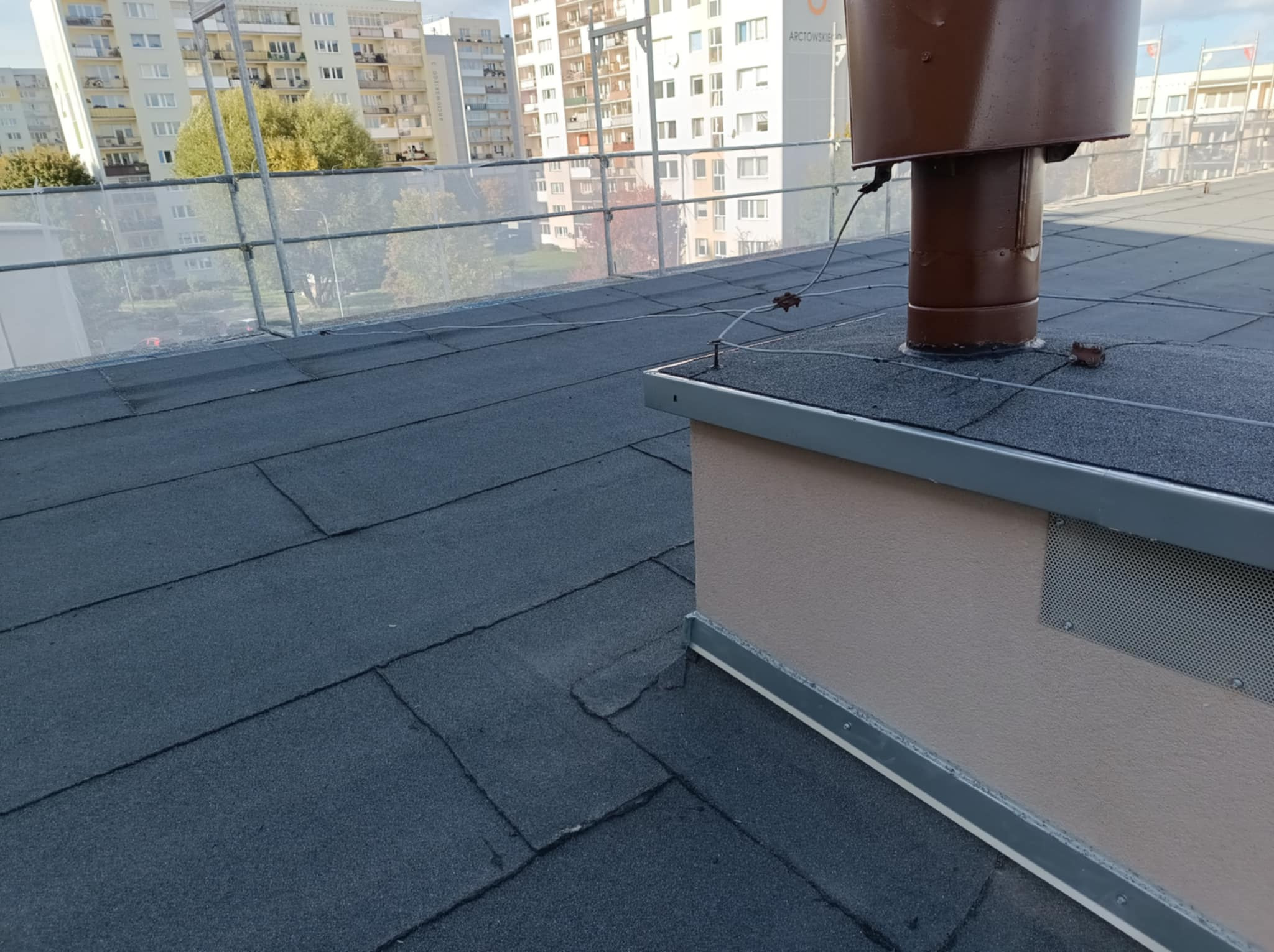 Płaski dach pokryty papą z brązowym kominem wentylacyjnym i zabezpieczeniami. W tle budynki mieszkalne i błękitne niebo. Widok z góry na fragment dachu.
