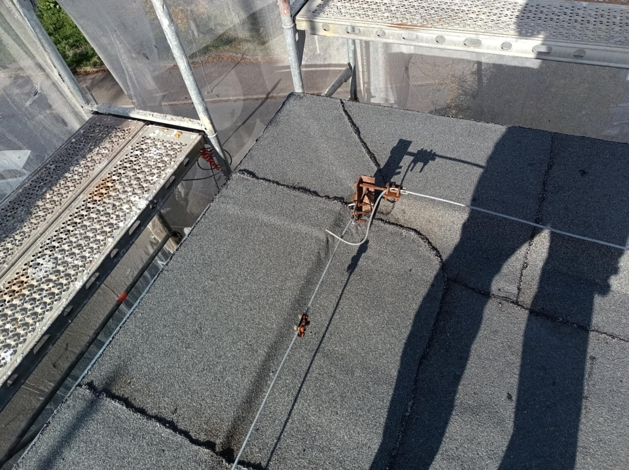 Szary dach z papy widziany z góry, z zamontowanym systemem odgromowym i fragmentem rusztowania. Widoczne łączenia papy i cień osoby wykonującej prace.