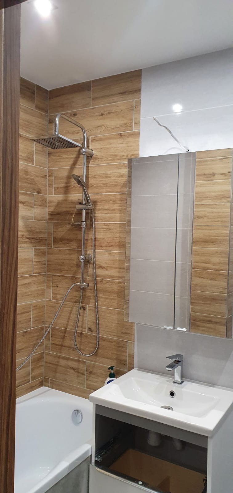 Ukończona łazienka z kabiną prysznicową typu walk-in, wykończona płytkami imitującymi drewno i szarą ceramiką, widoczna umywalka z szafką i lustrem.