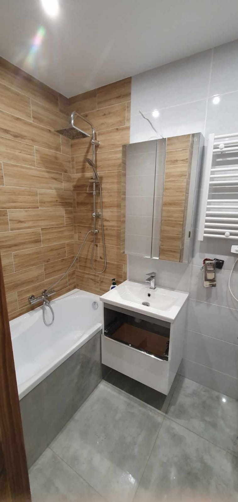 Nowoczesna łazienka po remoncie: drewnopodobne płytki na ścianie, biała wanna z szarą obudową, umywalka z szafką i lustrem, grzejnik drabinkowy.