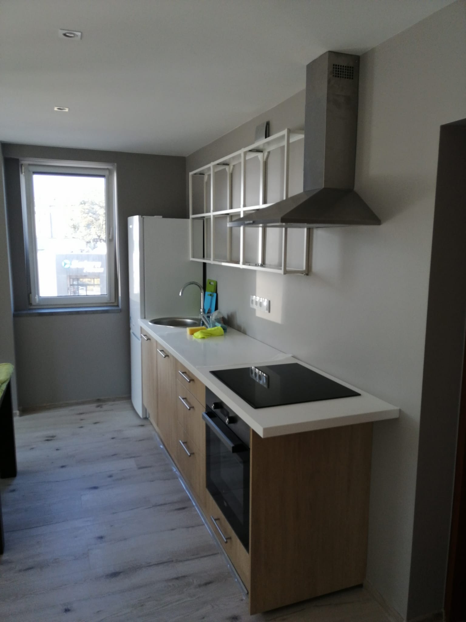 Nowoczesna kuchnia w szarościach z drewnianymi frontami szafek, wyposażona w płytę indukcyjną, okap, białą lodówkę i metalowe półki ścienne.