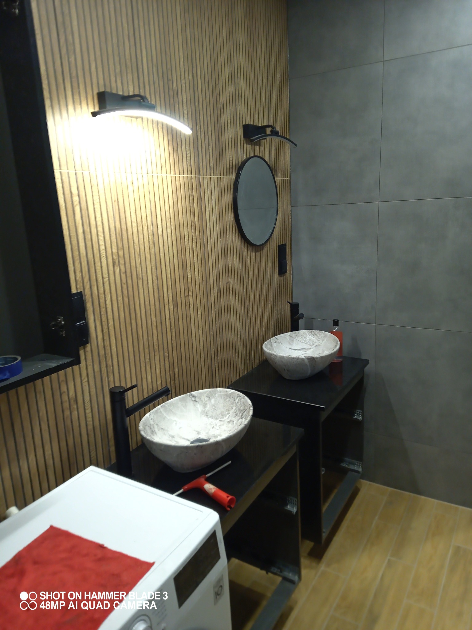 Nowoczesna łazienka z dwiema umywalkami nablatowymi o marmurkowym wzorze, czarną armaturą i szafkami, oświetleniem LED oraz drewnianą okładziną ścienną, z pralką i czerwonym materiałem w rogu kadru.