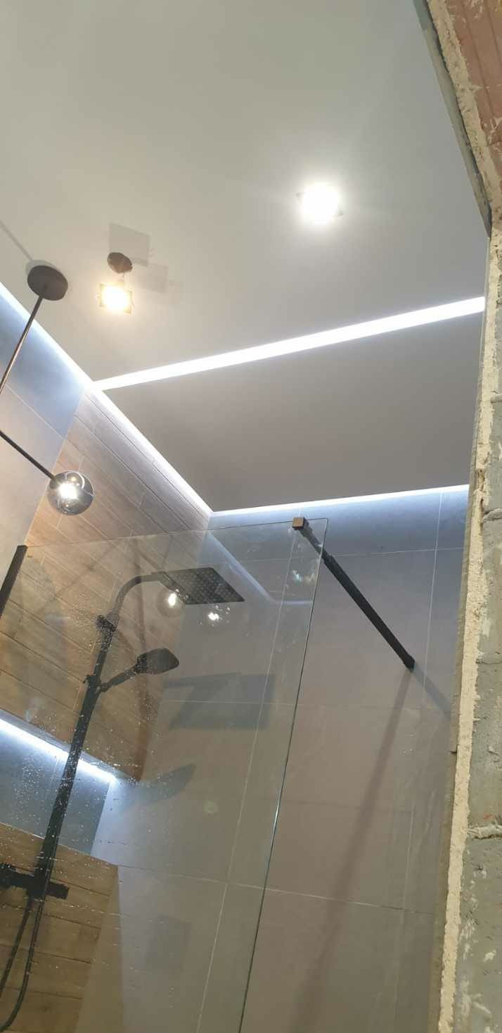 Nowoczesna kabina prysznicowa z czarną armaturą i oświetleniem LED, widok z dołu na sufit z dwoma rodzajami lamp i niedokończonym fragmentem ściany.