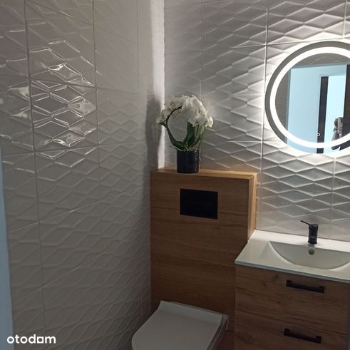 Nowoczesna łazienka z białymi trójwymiarowymi płytkami, podświetlanym okrągłym lustrem, drewnianą zabudową toalety i umywalki, oraz dekoracyjnym kwiatem w czarnym wazonie.