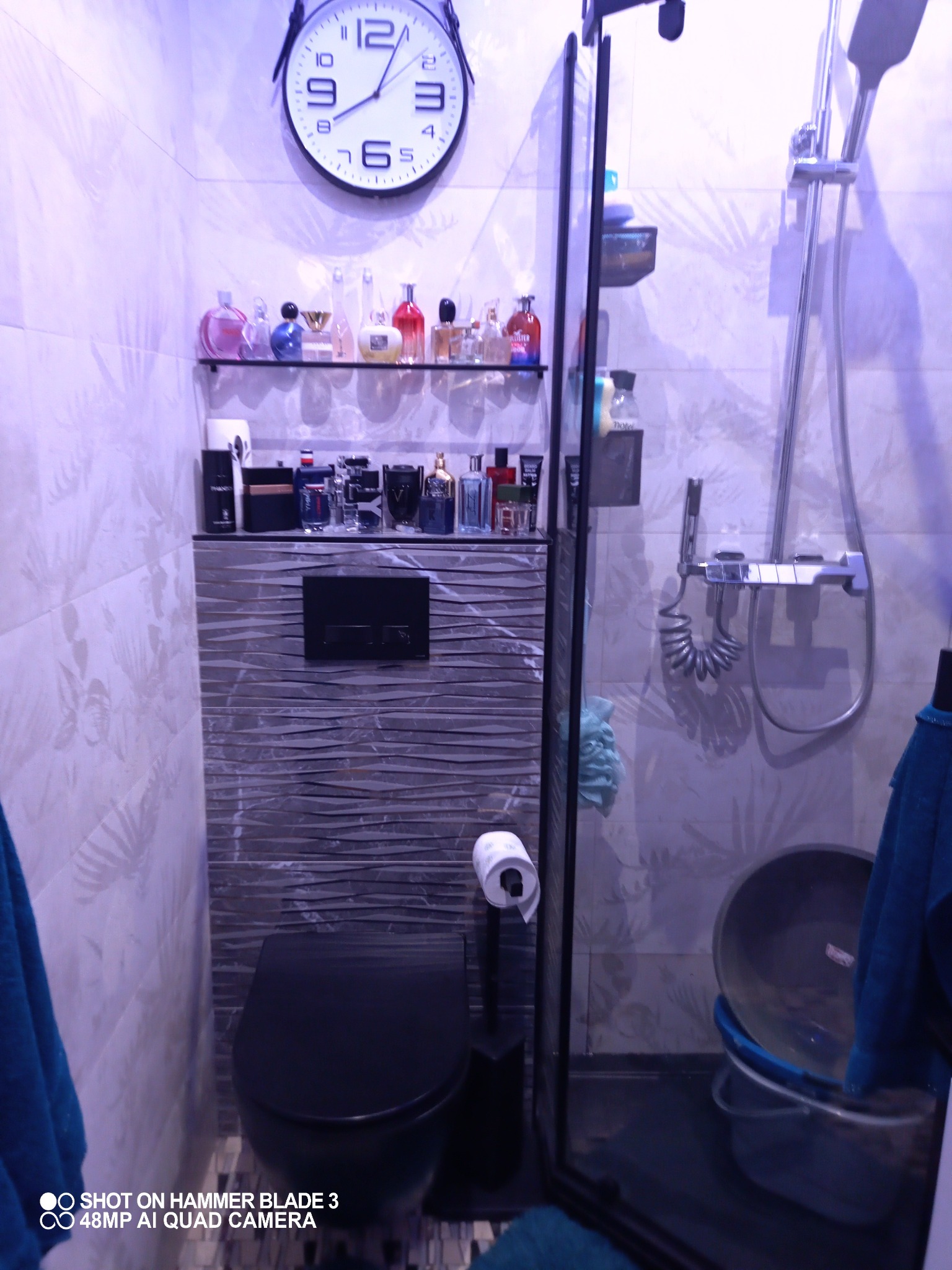 Nowoczesna łazienka z czarną toaletą, szklaną kabiną prysznicową i półką z perfumami, widoczny zegar na ścianie.