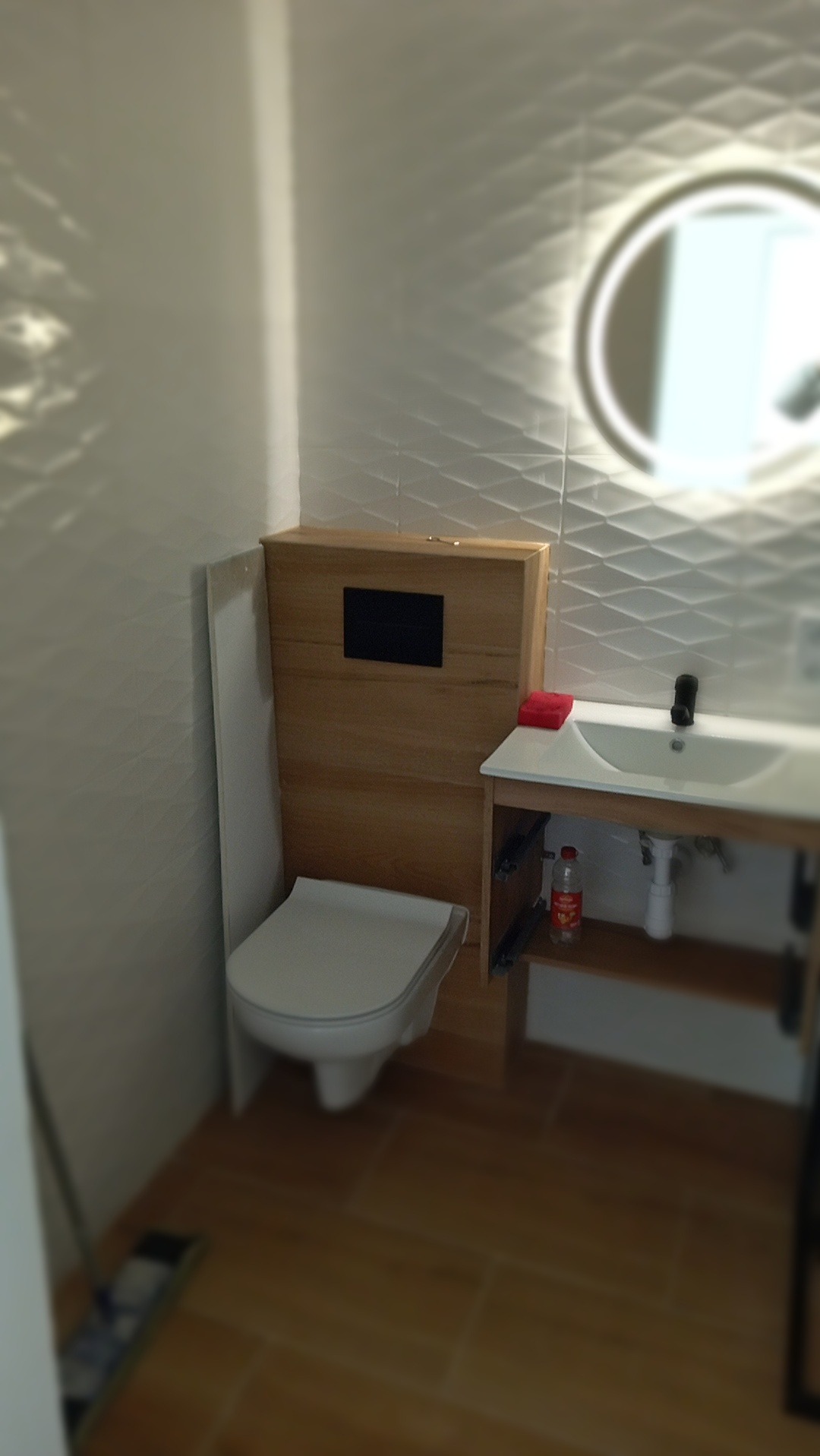 Nowoczesna łazienka z podwieszaną toaletą i umywalką z szafką w odcieniach drewna, oświetlone lustro na ścianie z trójwymiarową fakturą.