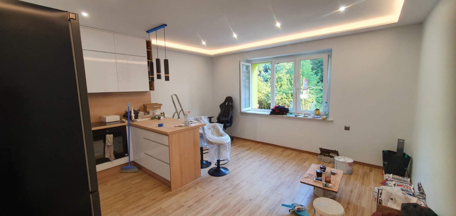 Wnętrze po remoncie: kuchnia z wyspą, białe szafki, drewniany blat, oświetlenie LED podsufitowe, krzesła zabezpieczone folią, widoczne ślady po pracach wykończeniowych.