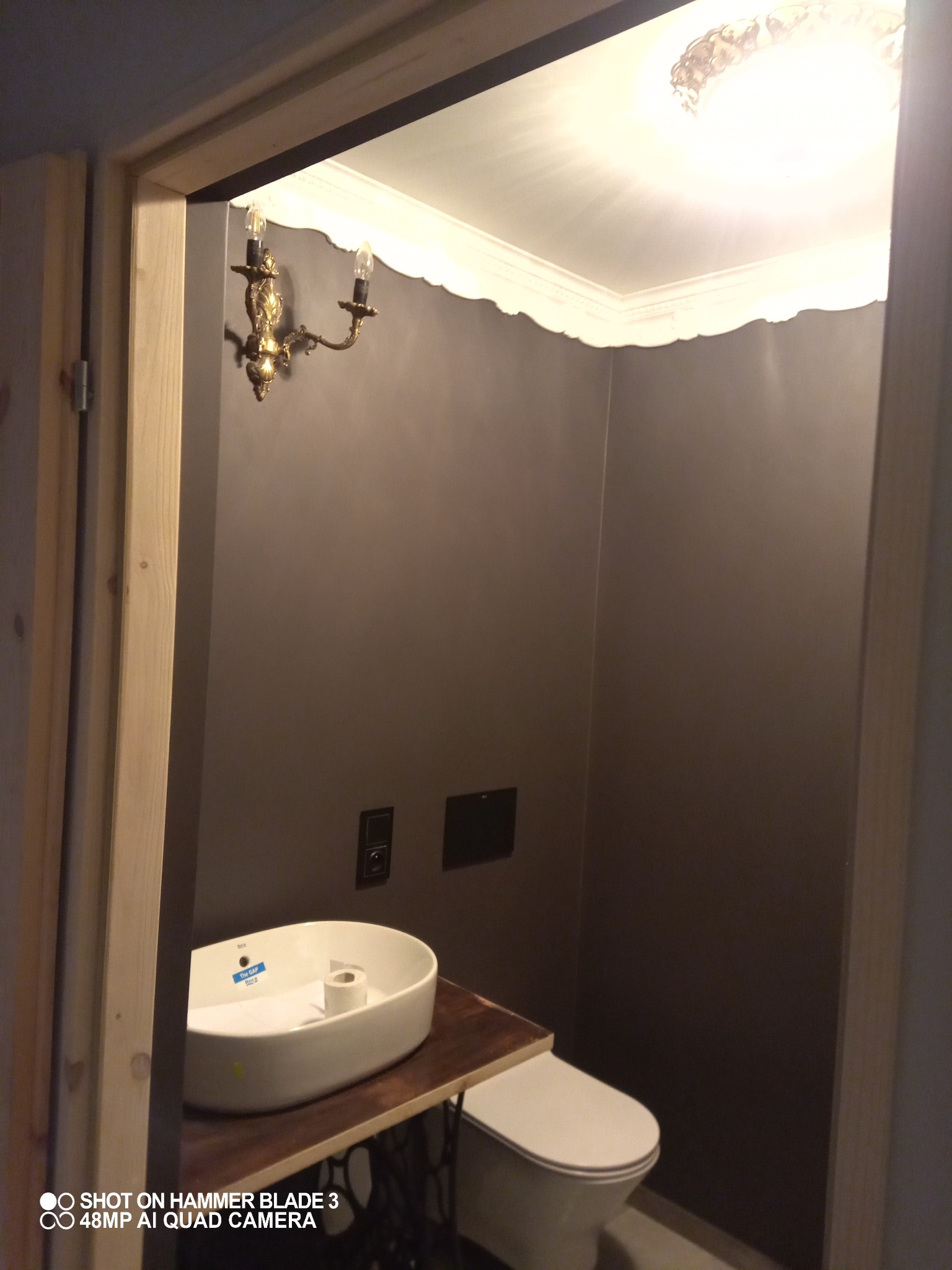 Mała łazienka w odcieniach szarości z umywalką na blacie z ciemnego drewna, wspartym na metalowej konstrukcji maszyny do szycia, oświetlona kinkietem w stylu retro i plafonem.