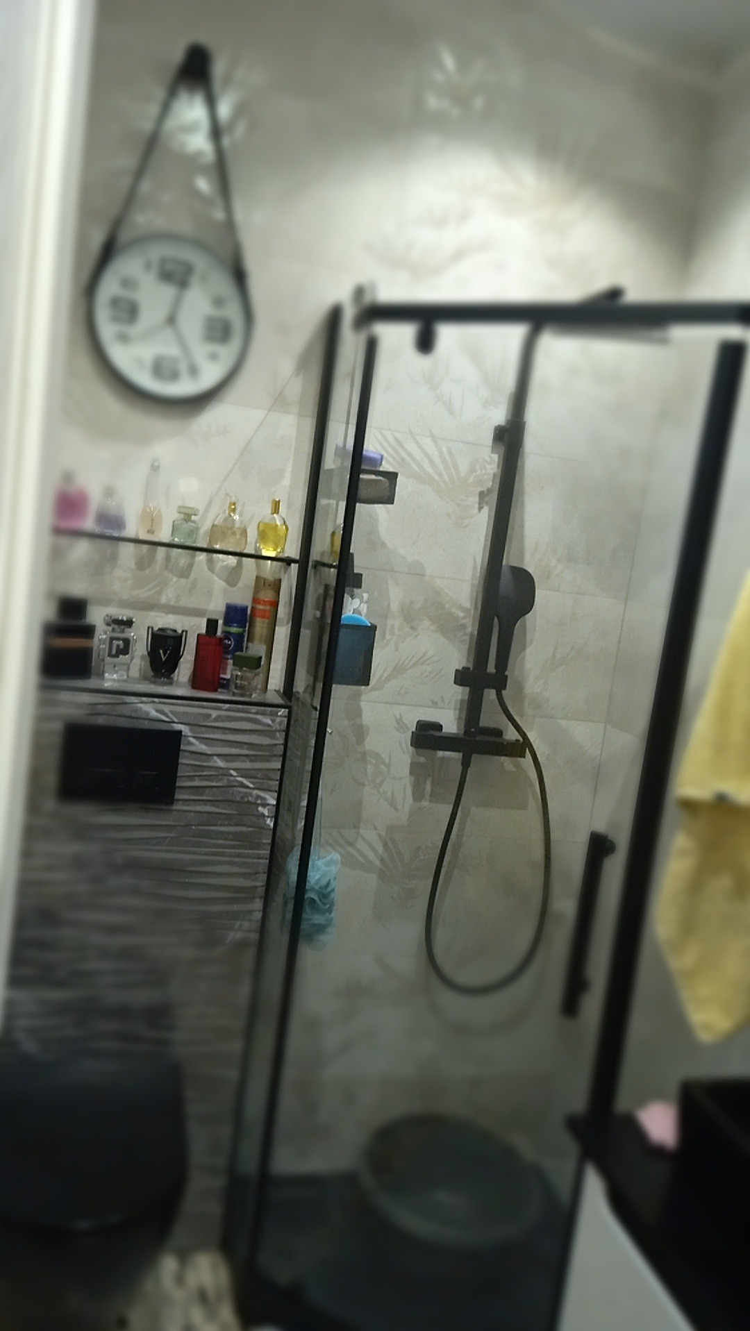 Nowoczesna kabina prysznicowa z czarnymi profilami, armaturą w tym samym kolorze i półką z kosmetykami, uzupełniona o zegar na ścianie.