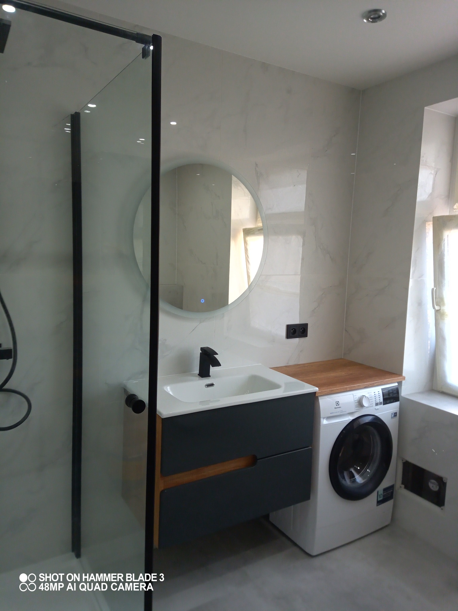 Nowoczesna łazienka z kabiną prysznicową typu walk-in, okrągłym lustrem z podświetleniem LED, umywalką z szafką w kolorze grafitowym z drewnianymi akcentami oraz pralką Electrolux z drewnianym...