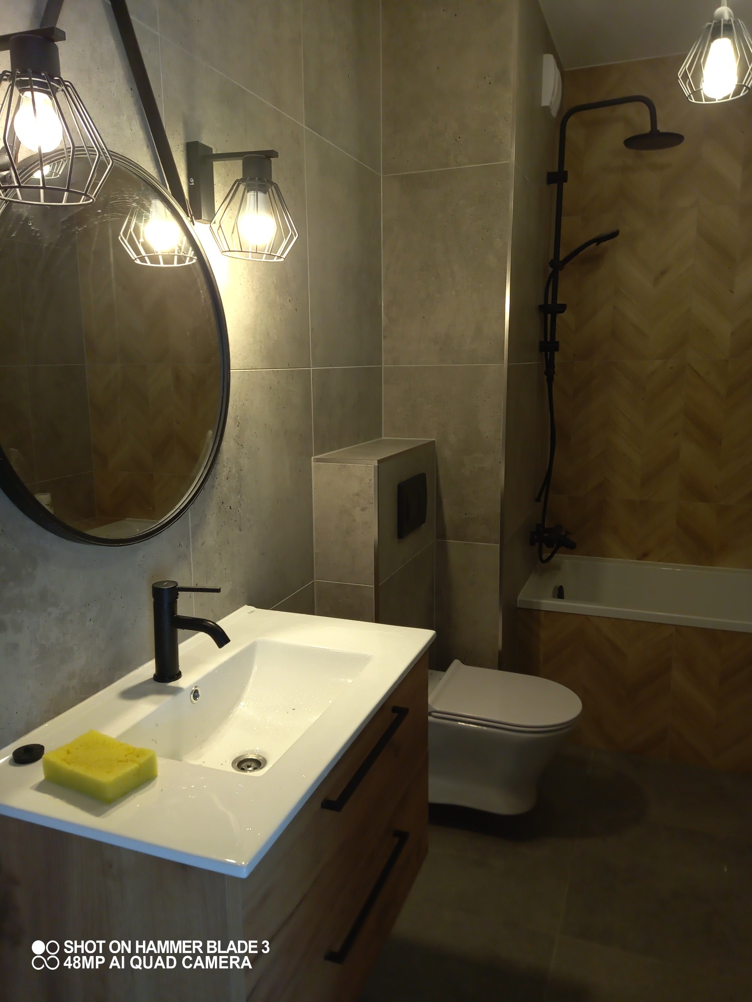 Wykończona łazienka z szarymi płytkami, drewnopodobną ścianą, wiszącą toaletą, umywalką z szafką i okrągłym lustrem z czarną ramą, oświetlona lampami z drucianymi kloszami, z gąbką leżącą na umywalce.