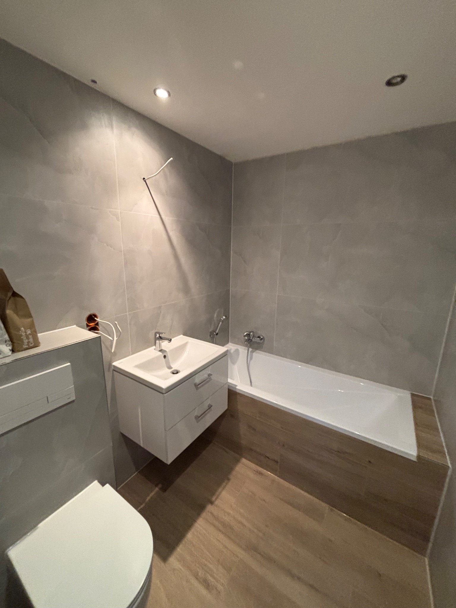 Nowoczesna łazienka z szarymi płytkami imitującymi marmur, podwieszaną białą szafką z umywalką, białą wanną z obudową z płytek drewnopodobnych i toaletą. Widoczne wystające przewody elektryczne...