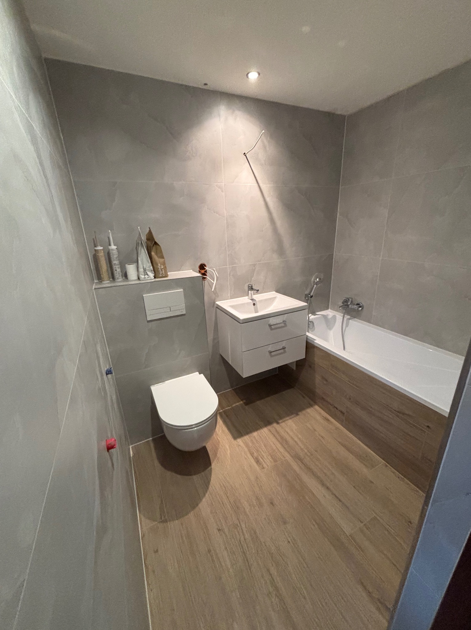 Nowoczesna łazienka z szarymi płytkami imitującymi kamień, drewnopodobną podłogą, podwieszaną toaletą i szafką z umywalką. Widoczne akcesoria łazienkowe i materiały budowlane na półce.