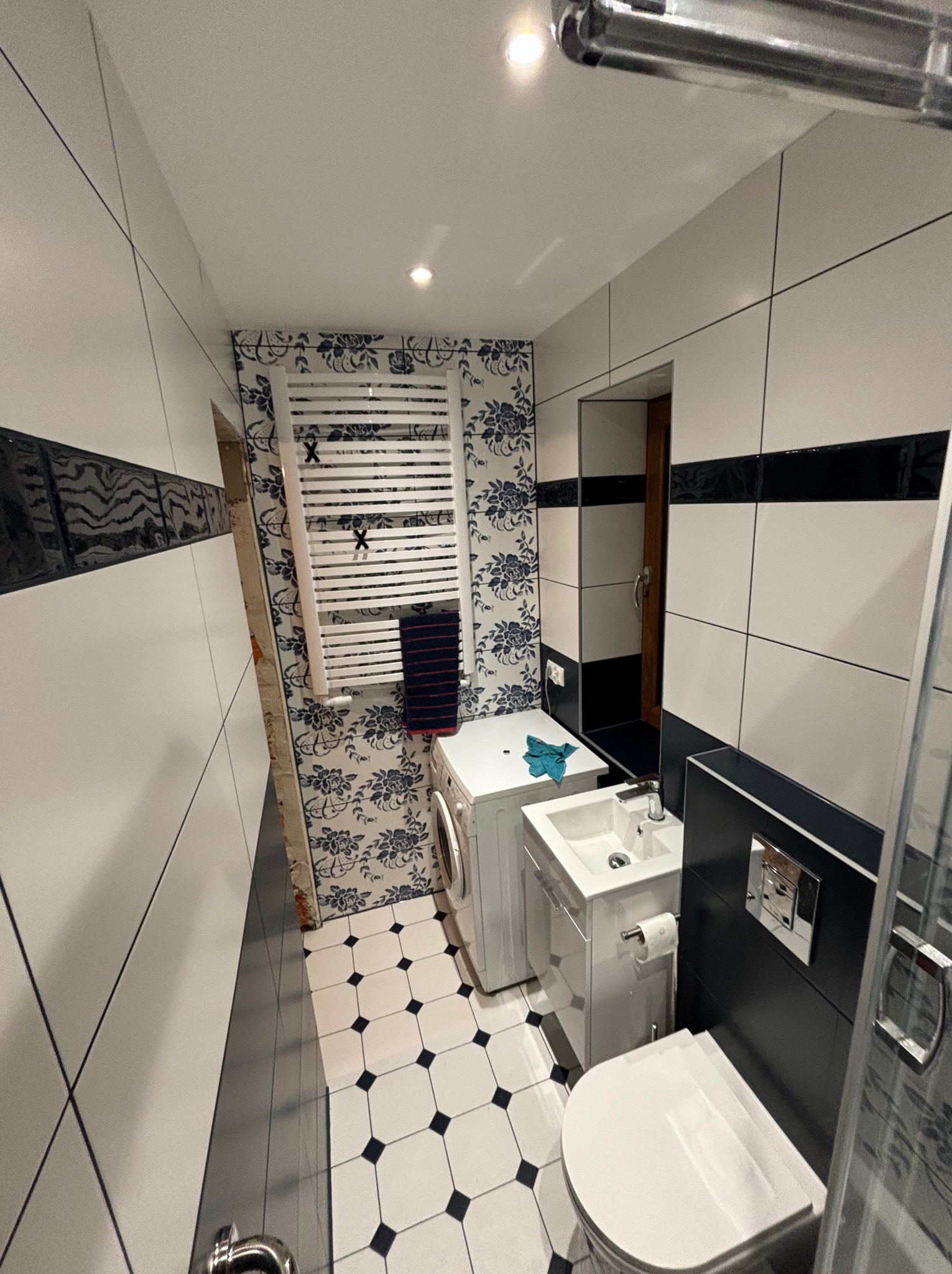 Wnętrze łazienki po remoncie: biało-czarne płytki podłogowe, ściany z białych płytek z czarnym pasem, dekoracyjna tapeta w kwiaty, biała pralka, umywalka z szafką, toaleta oraz grzejnik drabinkowy...
