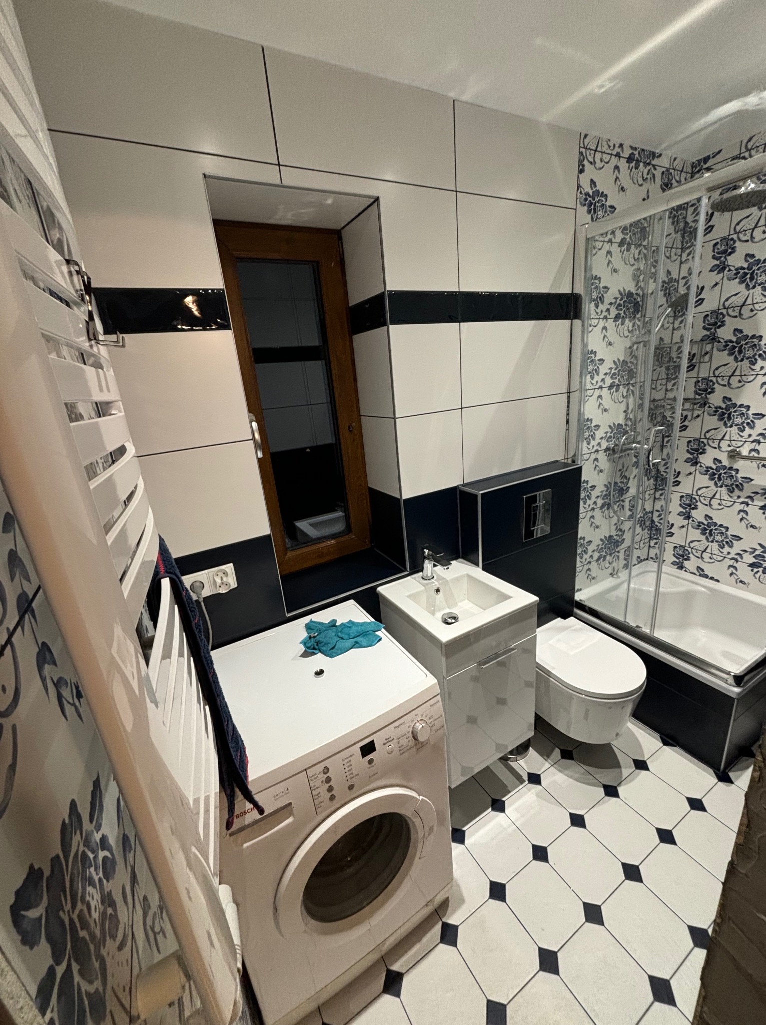 Wykończona łazienka z biało-czarnymi płytkami, kabiną prysznicową w kwiatowe wzory, umywalką z szafką, toaletą oraz pralką Bosch Serie 4, widoczny grzejnik drabinkowy.