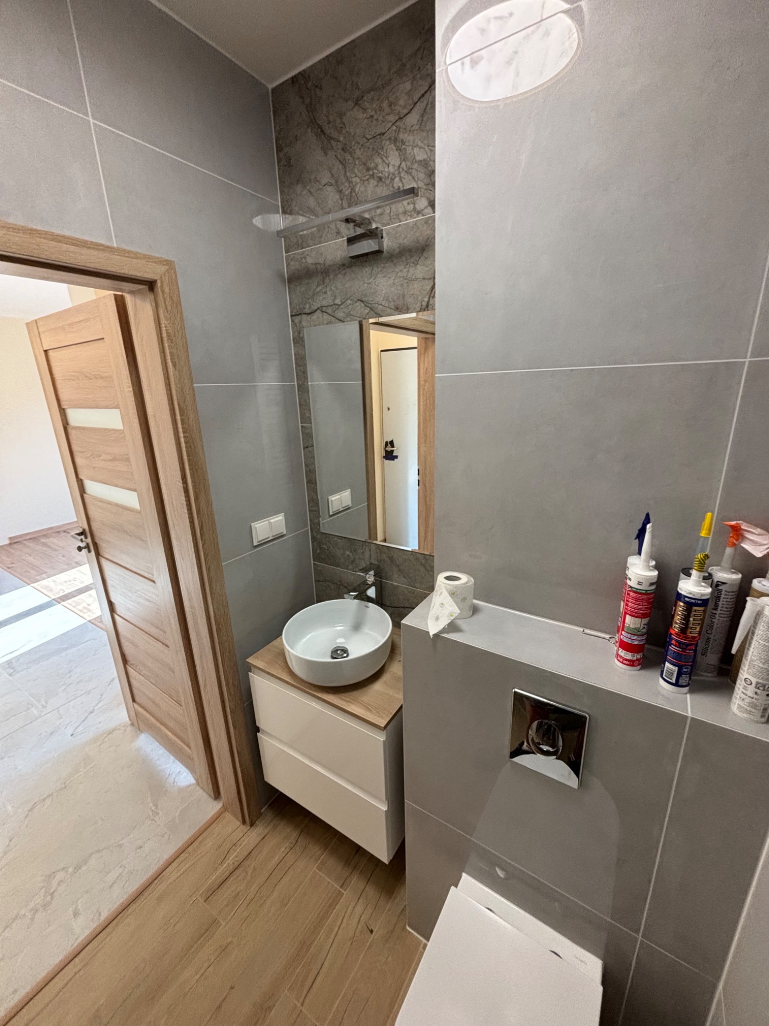 Nowoczesna łazienka z szarymi płytkami, okrągłą umywalką nablatową i drewnianą stolarką drzwiową; widoczne silikony i akryl na blacie.