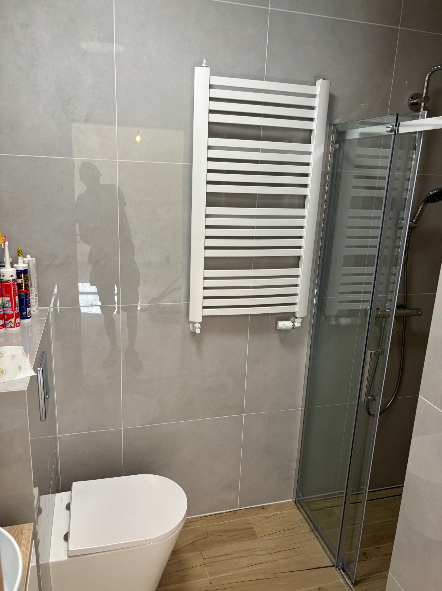 Nowoczesna łazienka z szarymi płytkami, białym grzejnikiem drabinkowym i kabiną prysznicową ze szklanymi drzwiami, widoczna toaleta i odbicie osoby w płytkach.