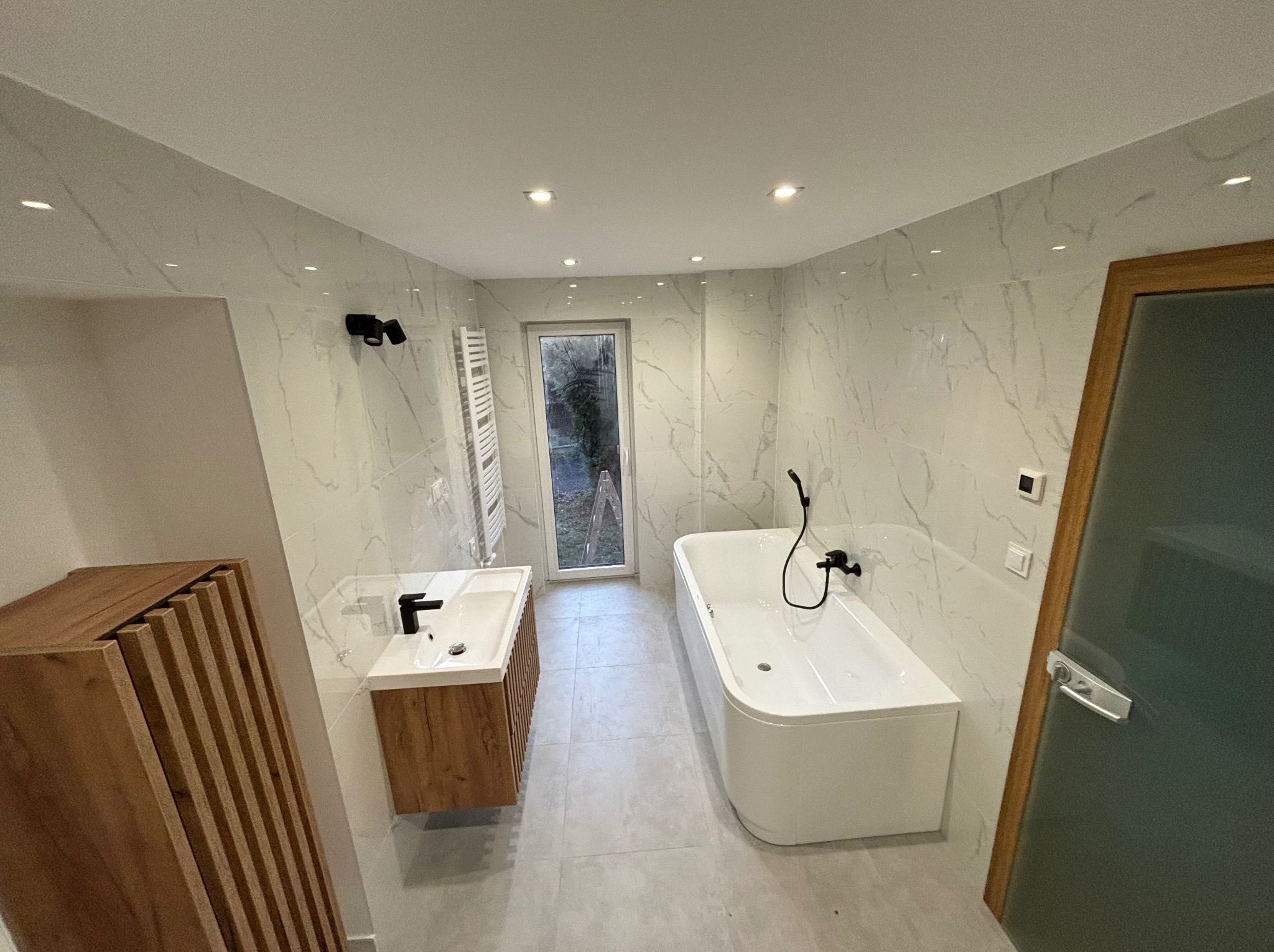 Nowoczesna łazienka z marmurowymi płytkami, drewnianą szafką pod umywalką z lamelami, białą wanną narożną i szarą podłogą z płytek, widok na okno z drabinką na ręczniki