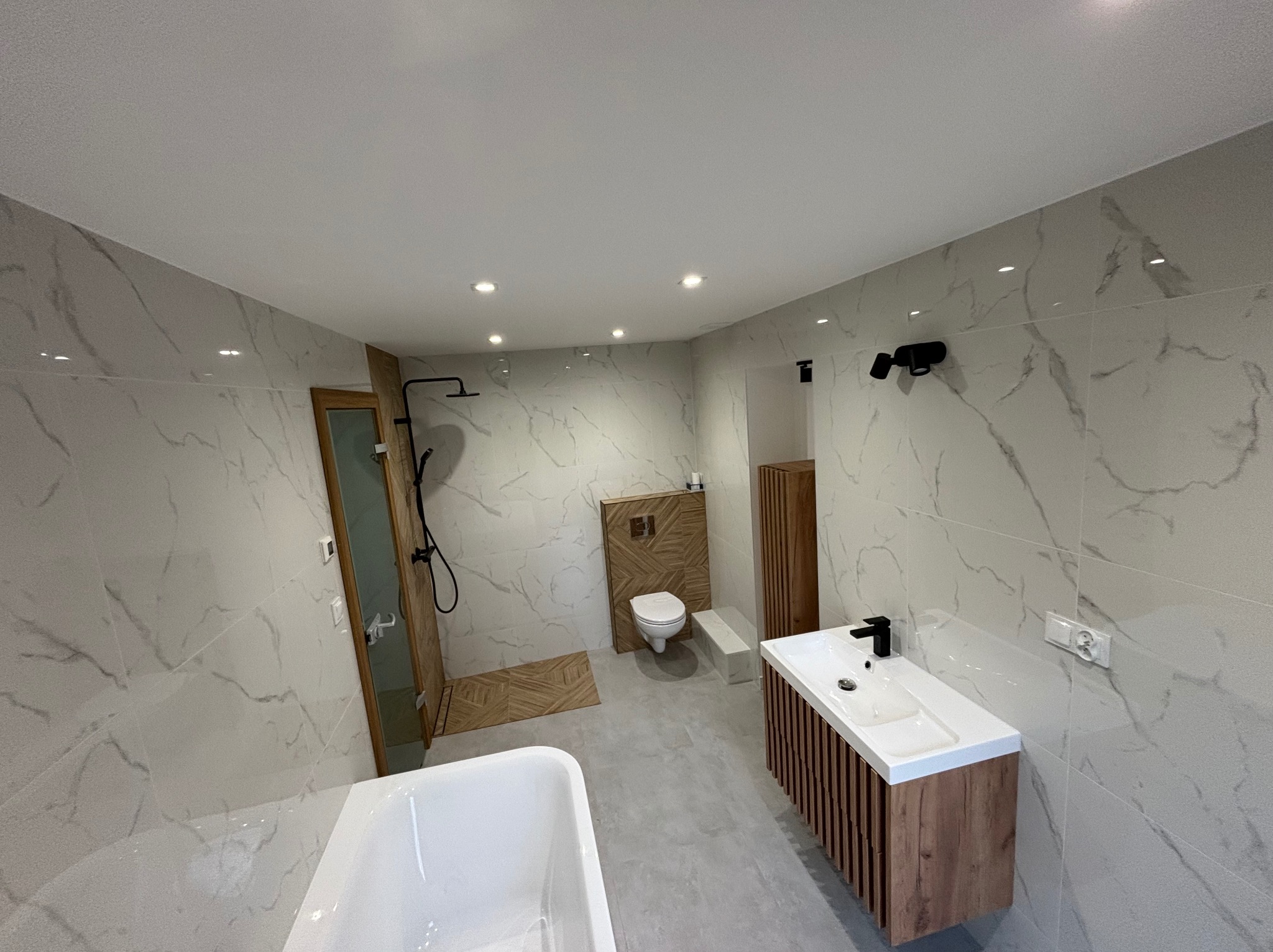 Nowoczesna łazienka z marmurowymi płytkami, drewnianymi akcentami i czarną armaturą, oświetlona punktowo, z widoczną wanną, umywalką z szafką i toaletą.