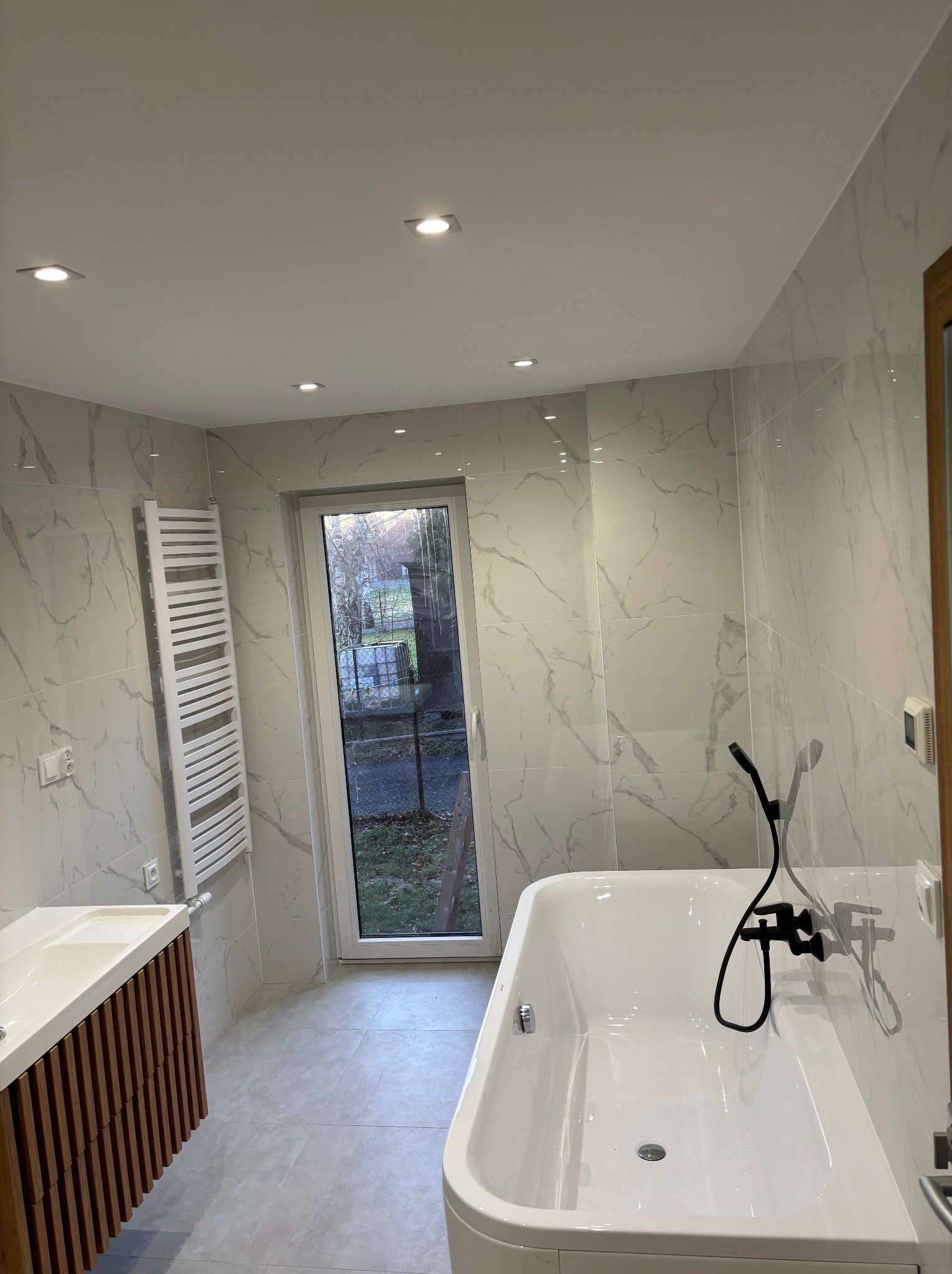 Nowoczesna łazienka z marmurowymi płytkami, białą wanną z czarną armaturą, umywalką z drewnianą szafką i grzejnikiem drabinkowym, oświetlona punktowymi lampami sufitowymi.