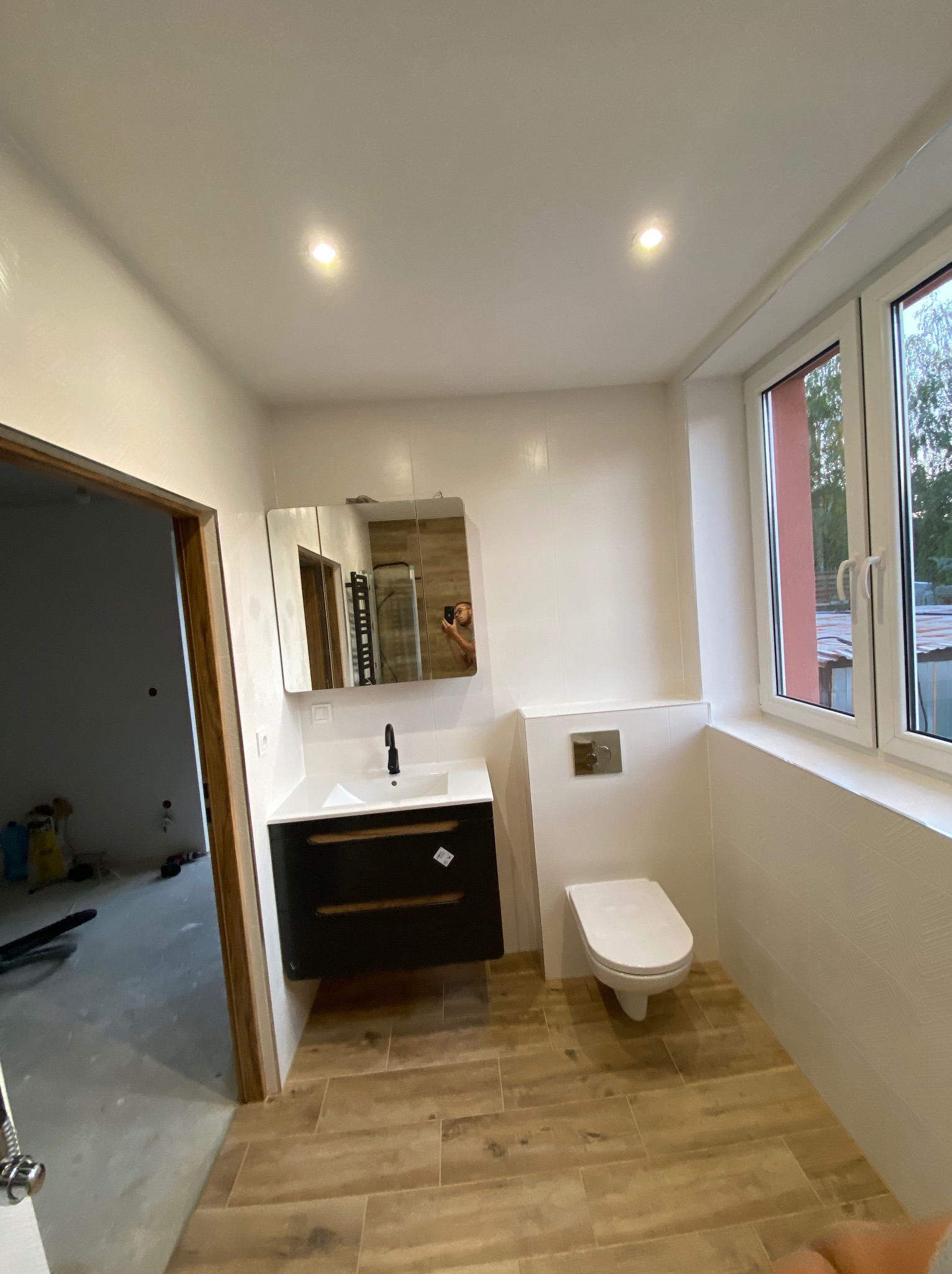 Nowoczesna łazienka z drewnopodobną podłogą, czarną szafką pod umywalką i białymi ścianami z teksturą, widok na okno i częściowo wykończone pomieszczenie obok.