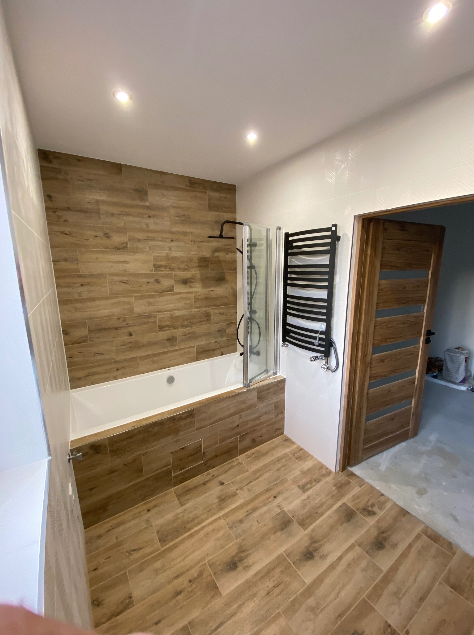 Nowoczesna łazienka z drewnopodobnymi płytkami na ścianach i podłodze, białą wanną z parawanem nawannowym, czarnym grzejnikiem drabinkowym i drewnianymi drzwiami.