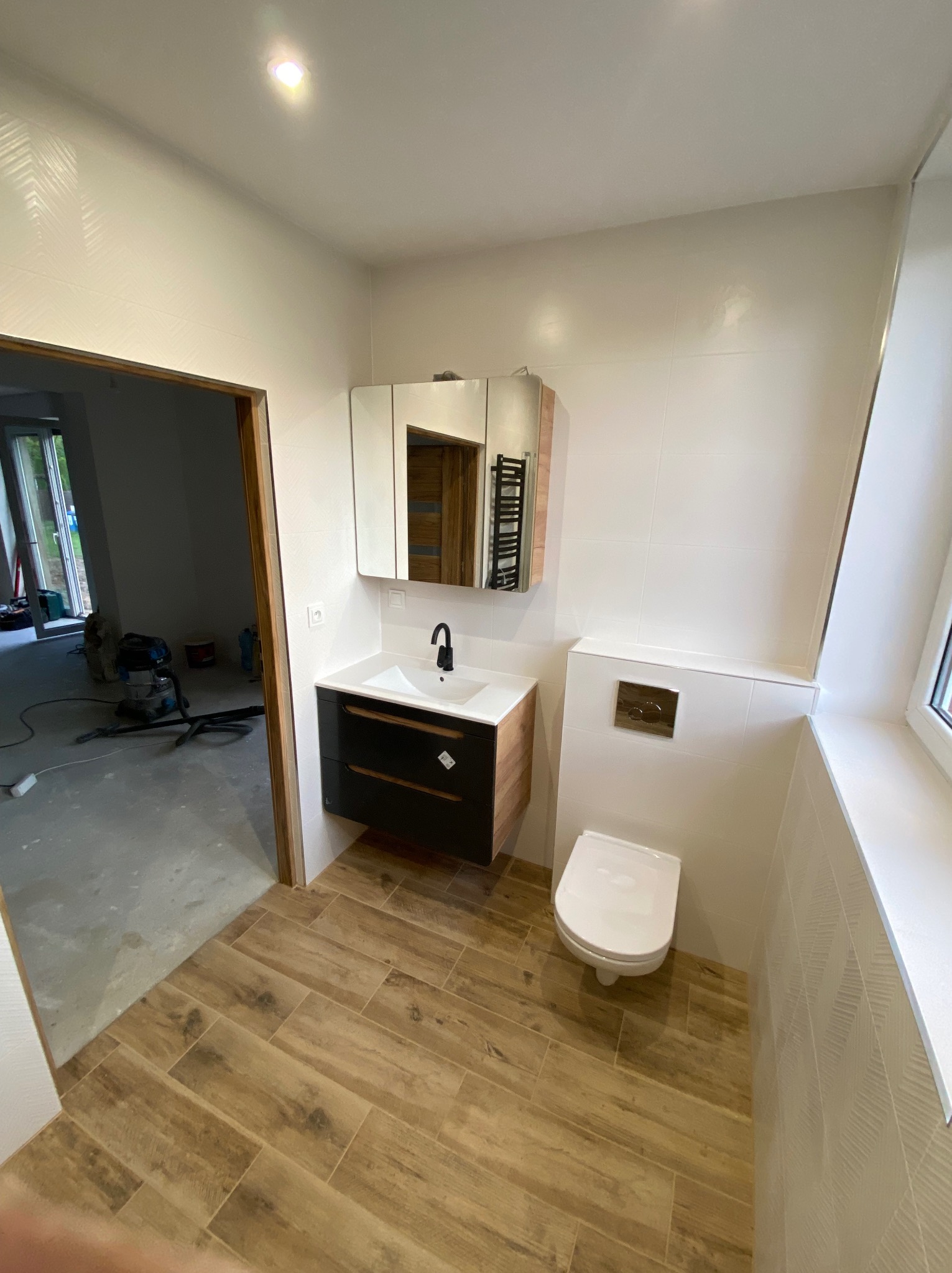 Nowoczesna łazienka z drewnopodobną podłogą, wiszącą toaletą i szafką z umywalką w kolorze drewna i czerni, widok na sąsiednie pomieszczenie w trakcie prac wykończeniowych.