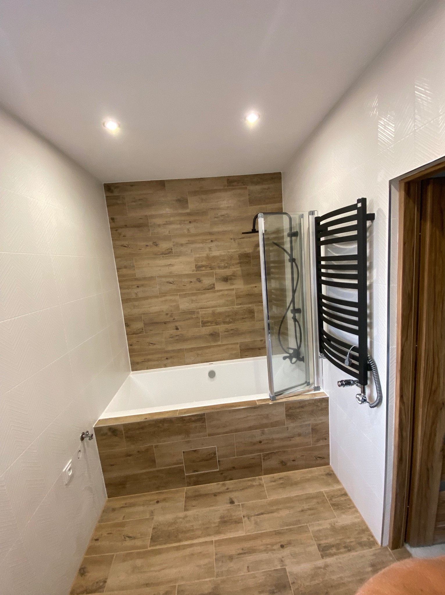 Nowoczesna łazienka w Bestwinie z wanną obudowaną płytkami imitującymi drewno, kabiną prysznicową z transparentnego szkła i czarnym grzejnikiem drabinkowym na tle białych ścian z delikatnym wzorem.