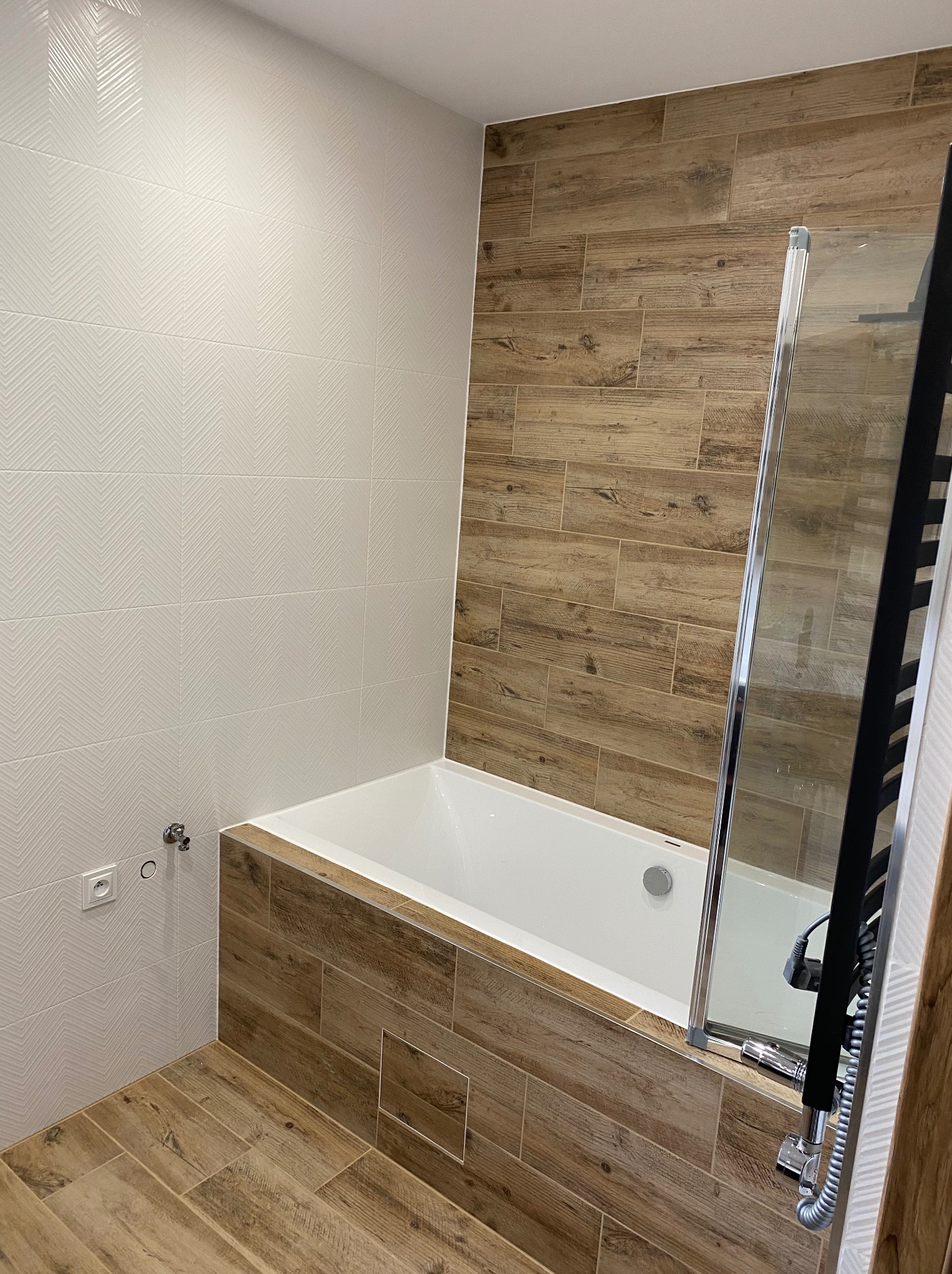 Nowoczesna łazienka z wanną obudowaną płytkami imitującymi drewno, kabina prysznicowa ze srebrnymi profilami, ściany wyłożone białymi płytkami z delikatnym wzorem jodełki, drewnopodobna podłoga...