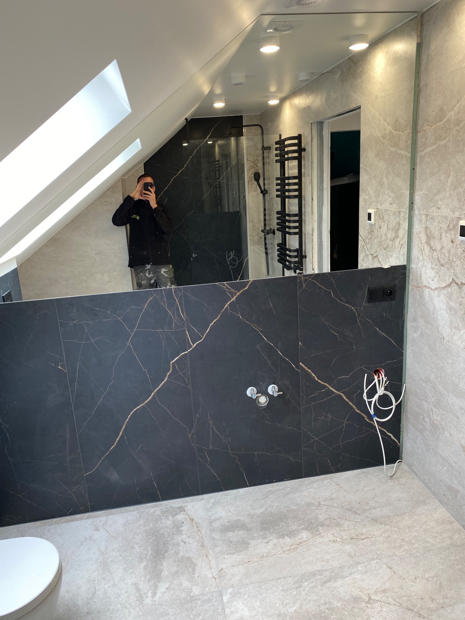 Nowoczesna łazienka na poddaszu z czarną marmurową ścianą, widoczne przyłącza hydrauliczne i elektryczne, odbicie fotografa w dużym lustrze.