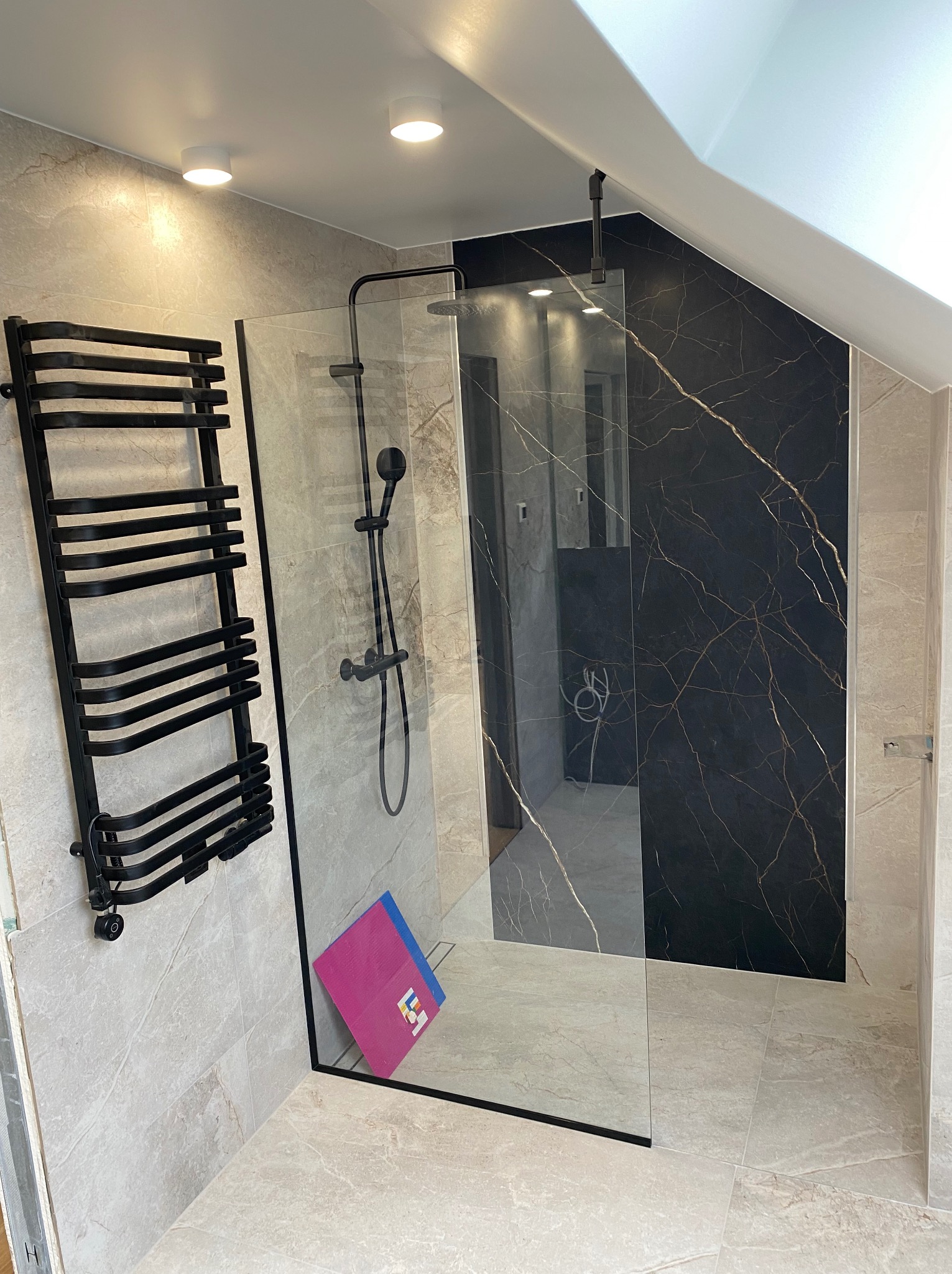 Wykończona łazienka na poddaszu ze ścianą prysznicową z czarnymi elementami, grzejnikiem drabinkowym i płytkami imitującymi marmur.